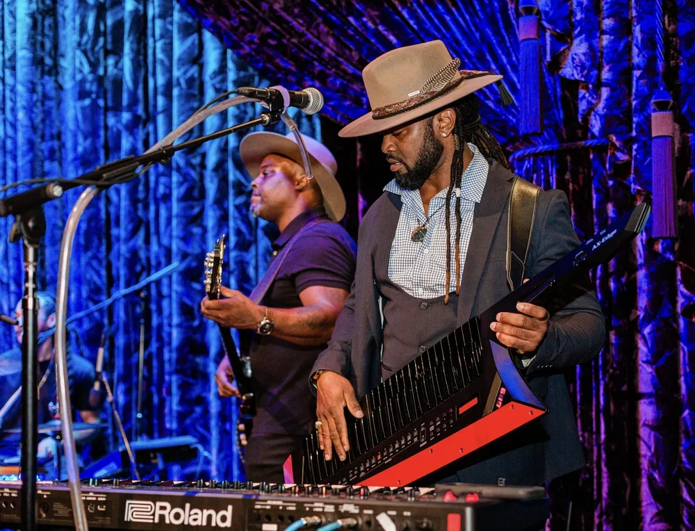 Two musicians perform on stage with purple and blue lighting. One is playing a keytar, and the other is playing a guitar. Both are wearing hats and stylish clothing.