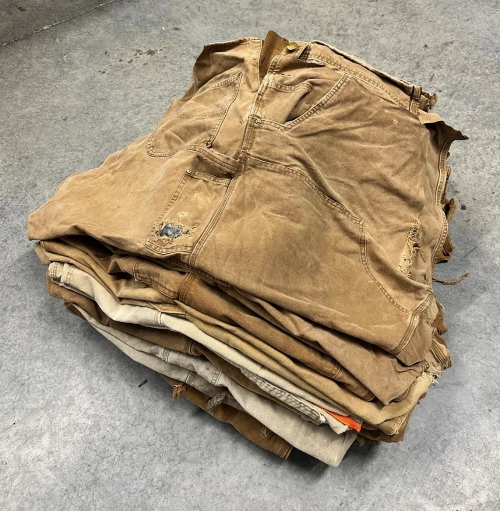 A pile of worn and stacked tan and beige jeans on a concrete floor.