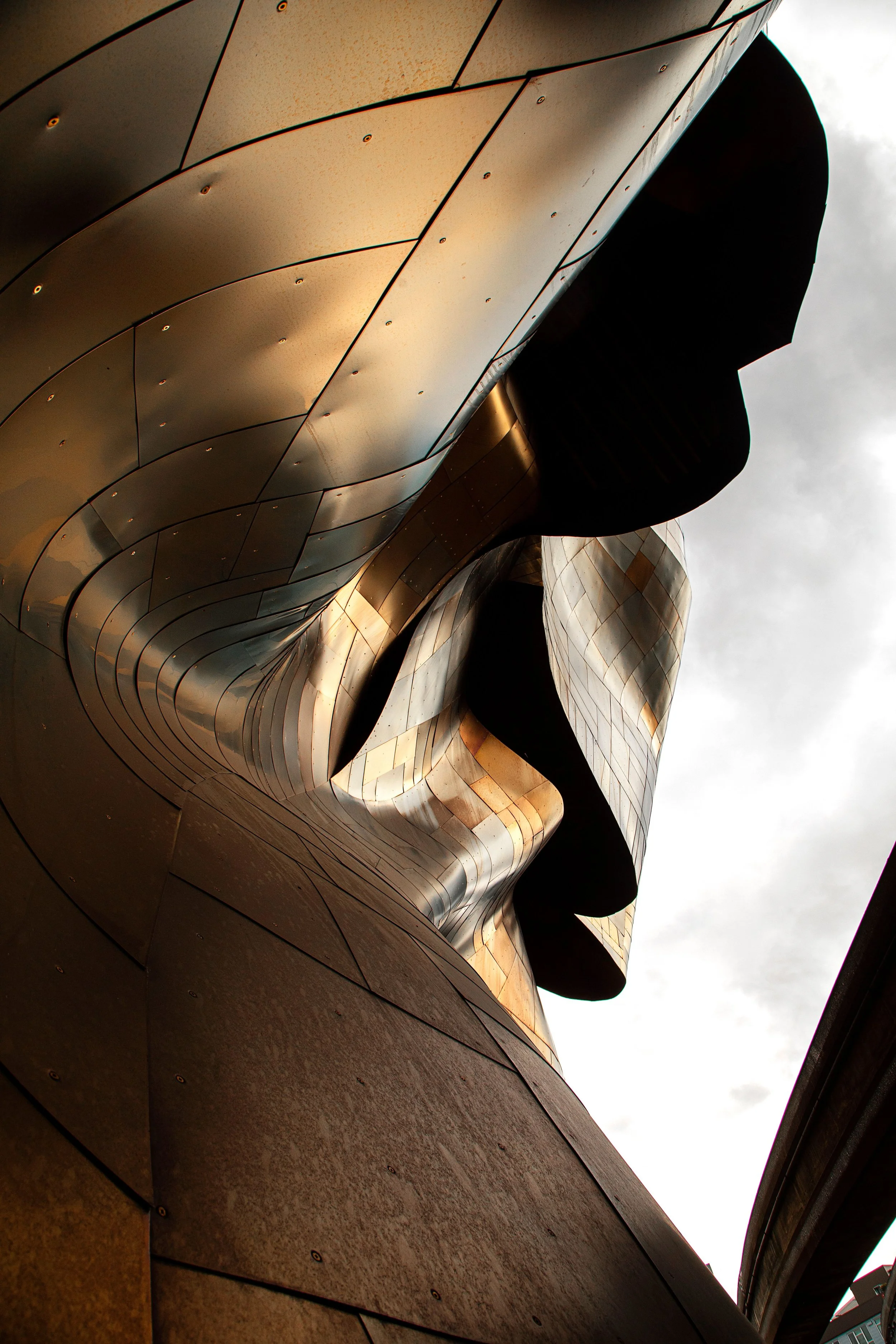 Close-up of a modern architectural building with curving metallic panels and a human face-shaped sculpture integrated into the design, with a cloudy sky background.