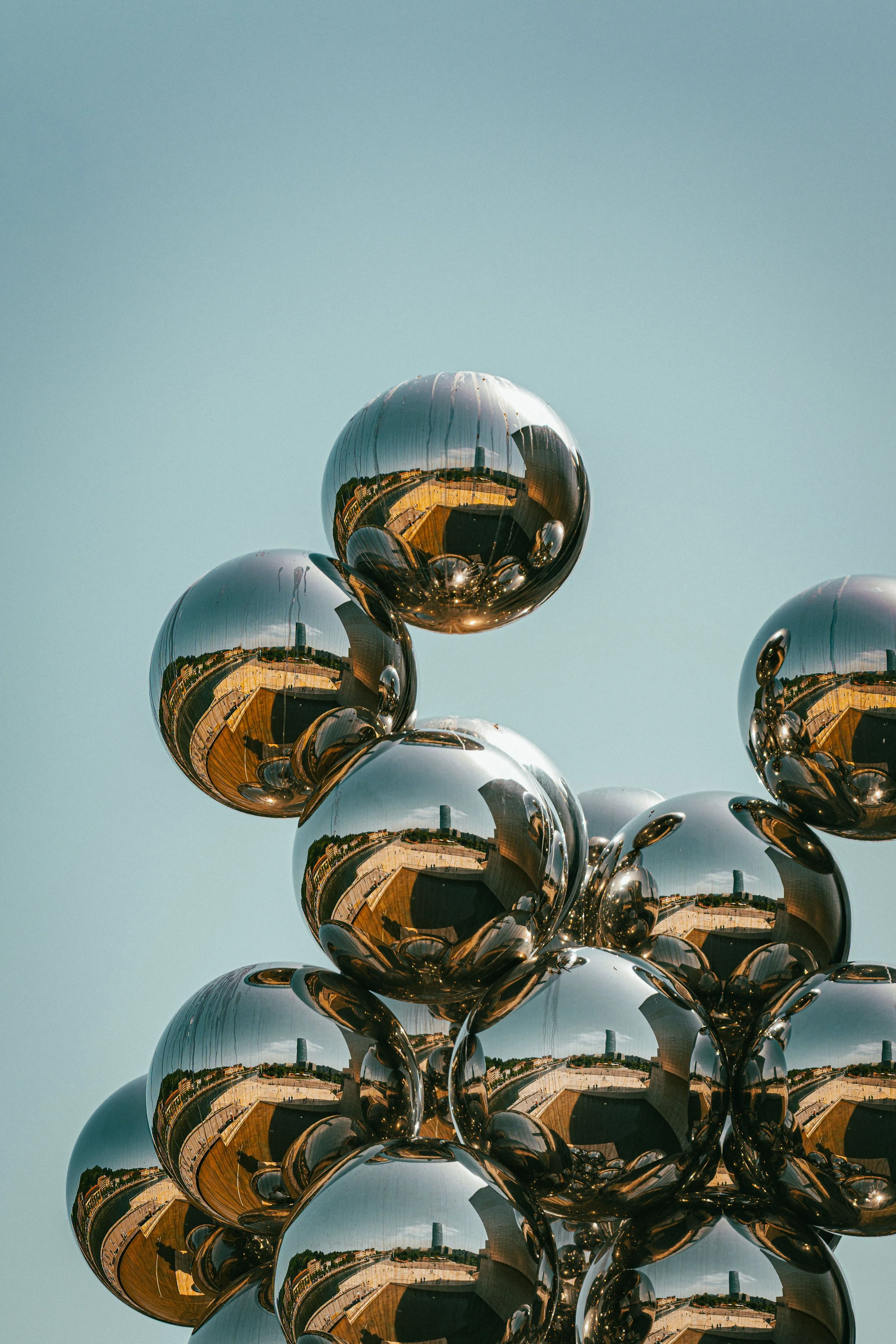 Reflective metallic spheres stacked in a pyramid formation outdoors, with a cityscape and blue sky reflected in their surfaces.