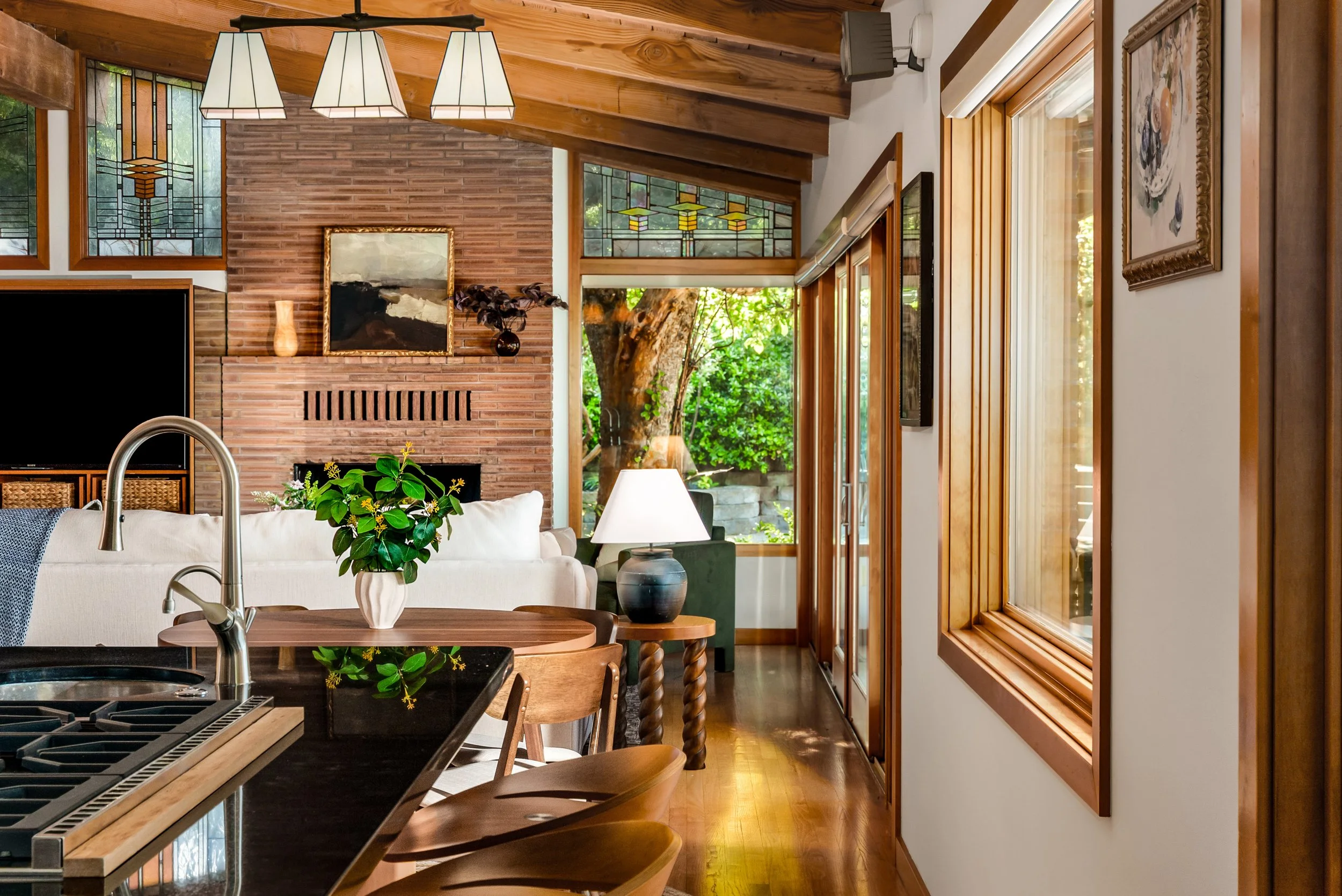 Inside view of a living room with wooden ceiling beams, stained glass windows, brick fireplace, and large windows showing green outdoor scenery. Contains sofa, table, plants, and artwork.