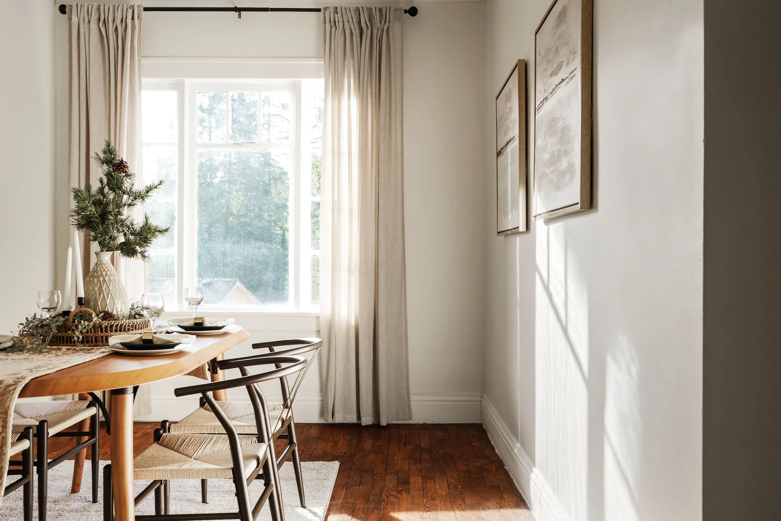 A dining room with a wooden table set for a meal, decorated with a woven basket, greenery, and wine glasses. A large window with sheer curtains and beige drapes lets in natural light. The walls are decorated with abstract artwork, and there is a smal