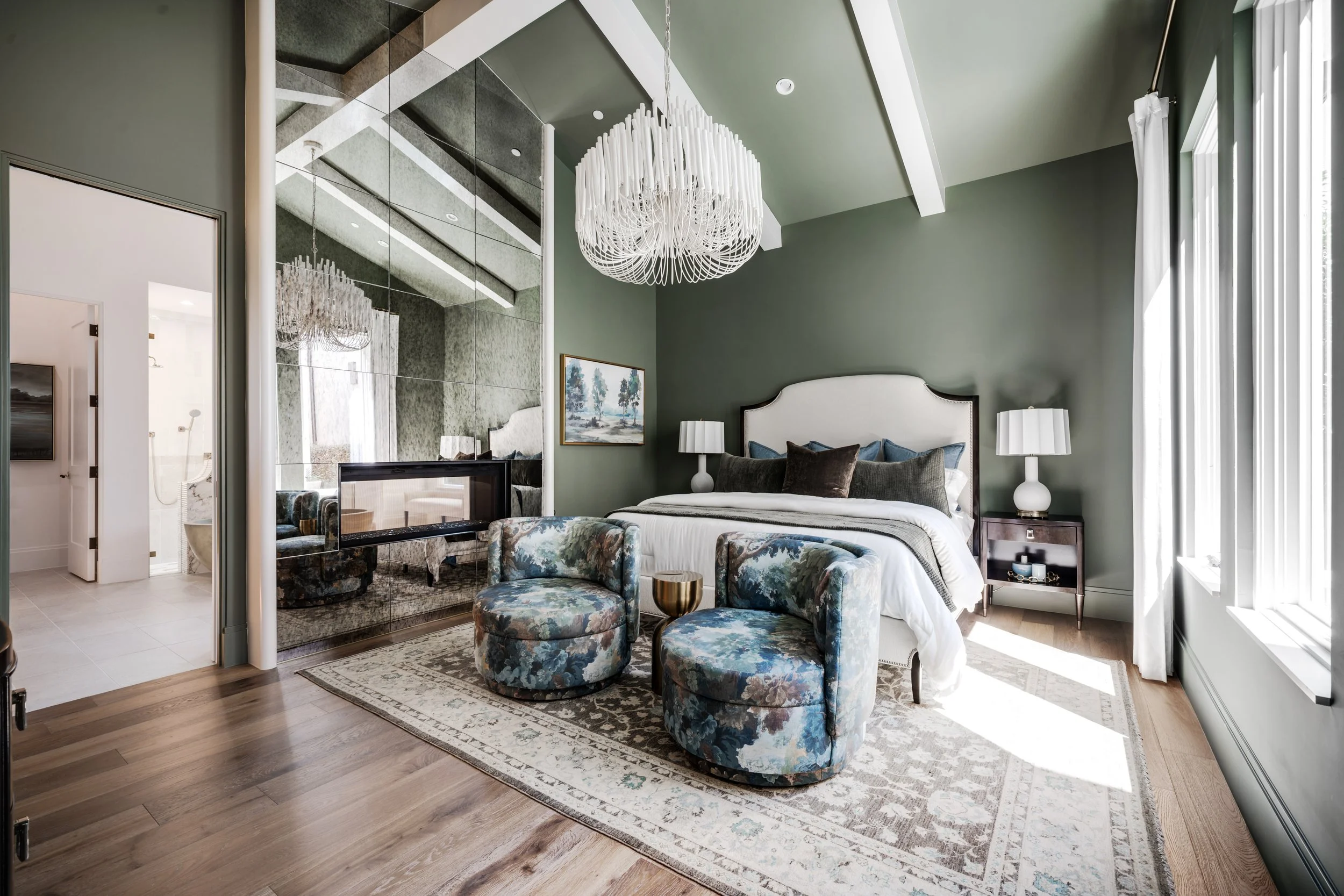 A bedroom with green walls, large window with white curtains, white bed with pillows, two white nightstands with lamps, a mirror on the wall, and a decorative chandelier hanging from the ceiling. There are mirrored sliding closet doors and a painting