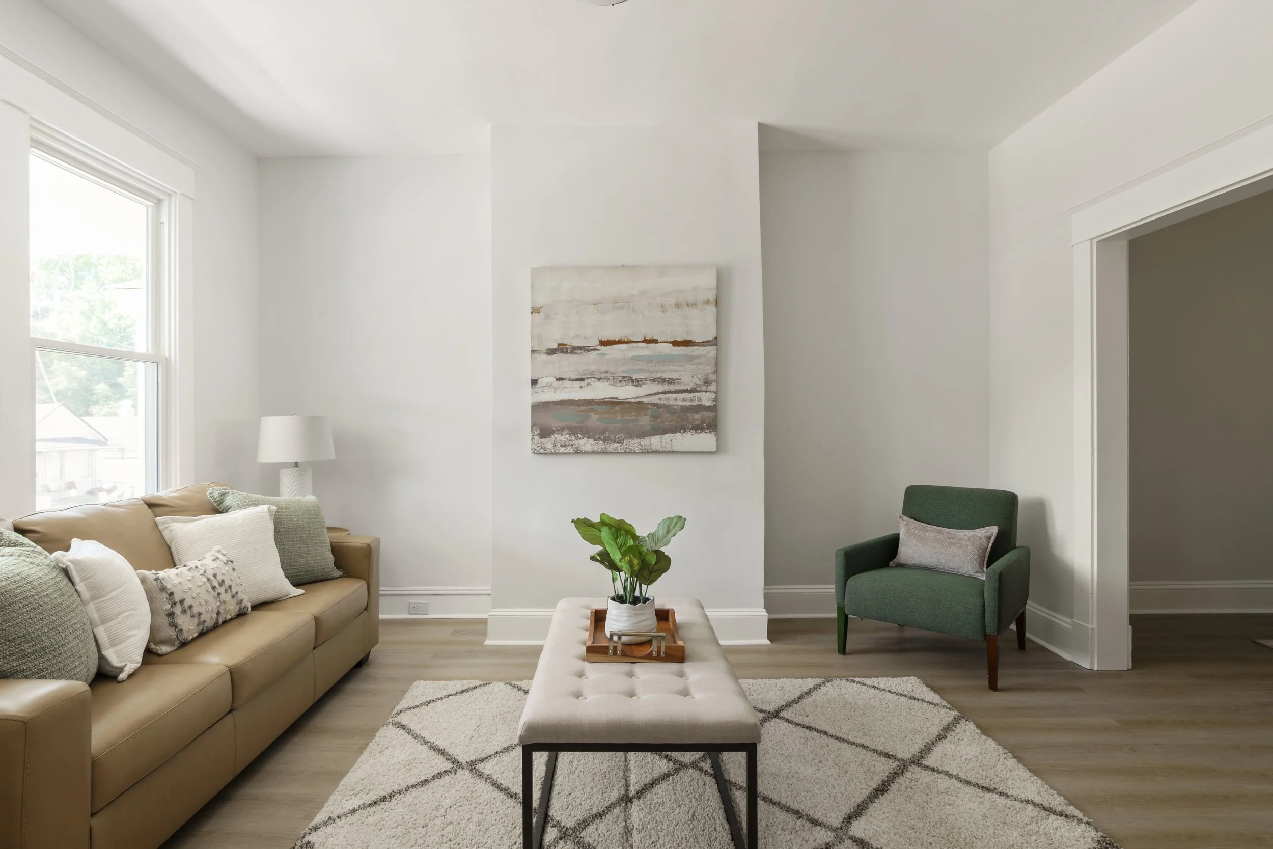 Living room with a beige sofa, green armchair, white ottoman with a plant, abstract painting, and large window in natural light.