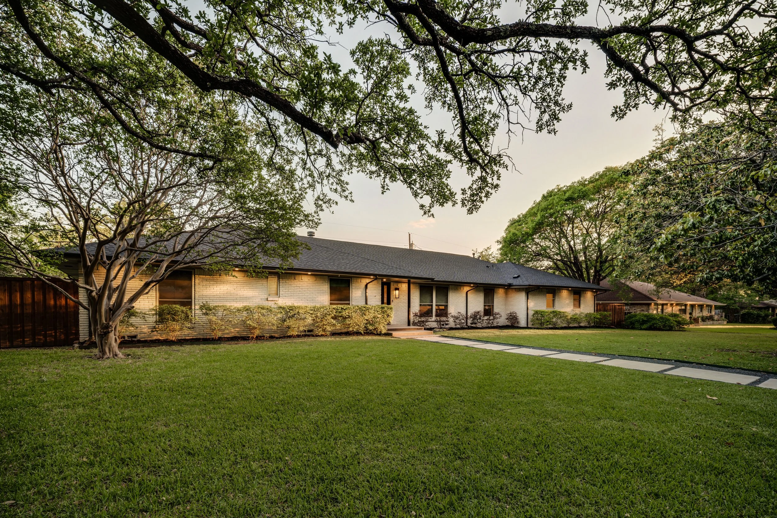 Backyard with lawn, walkway, trees, and a single-story house with a dark roof in the background during early evening or late afternoon.