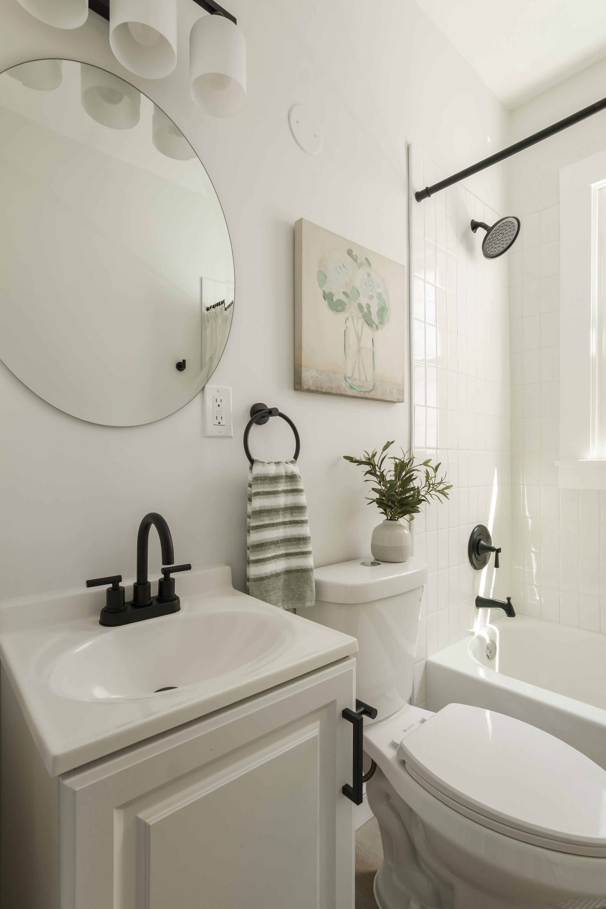 A modern bathroom with a white vanity, black faucet, large round mirror, and white wall tiles. The shower features a black rain showerhead and a black shower handle. There is a small art piece on the wall with a vase of flowers, a potted plant on the