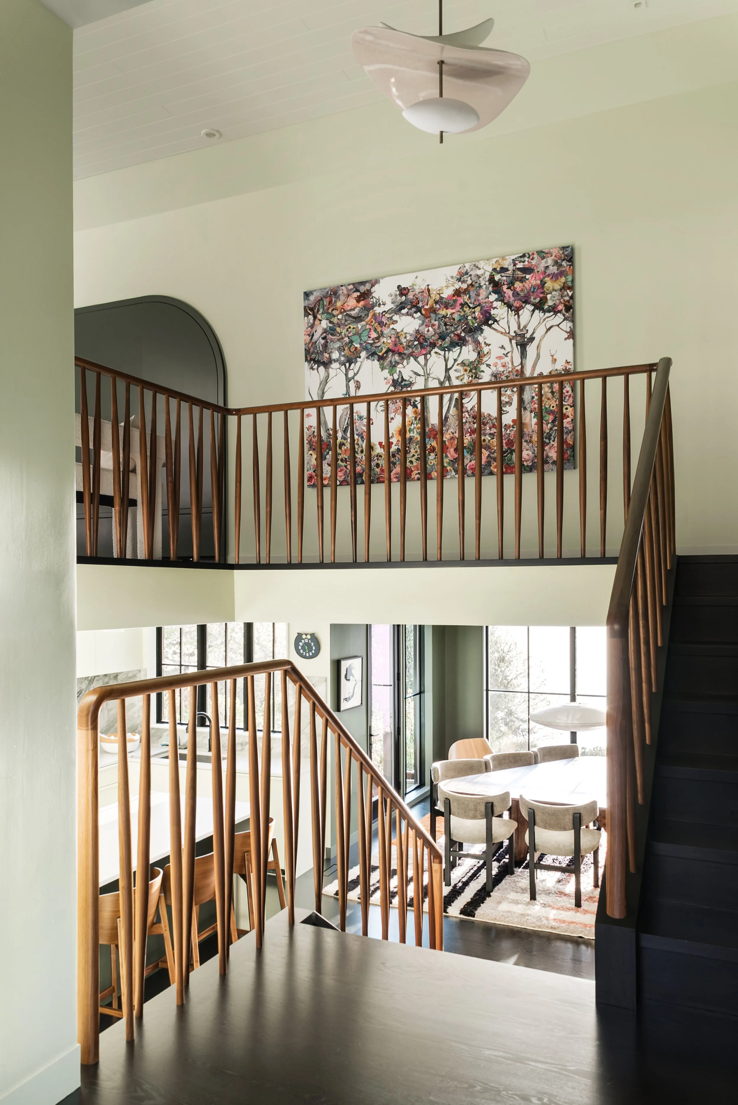 A two-story open living space with a staircase and a railing. The upper level has abstract floral artwork on a wall, while the lower level features large windows, a dining area with chairs, and a living room with a circular rug and seating. The ceili