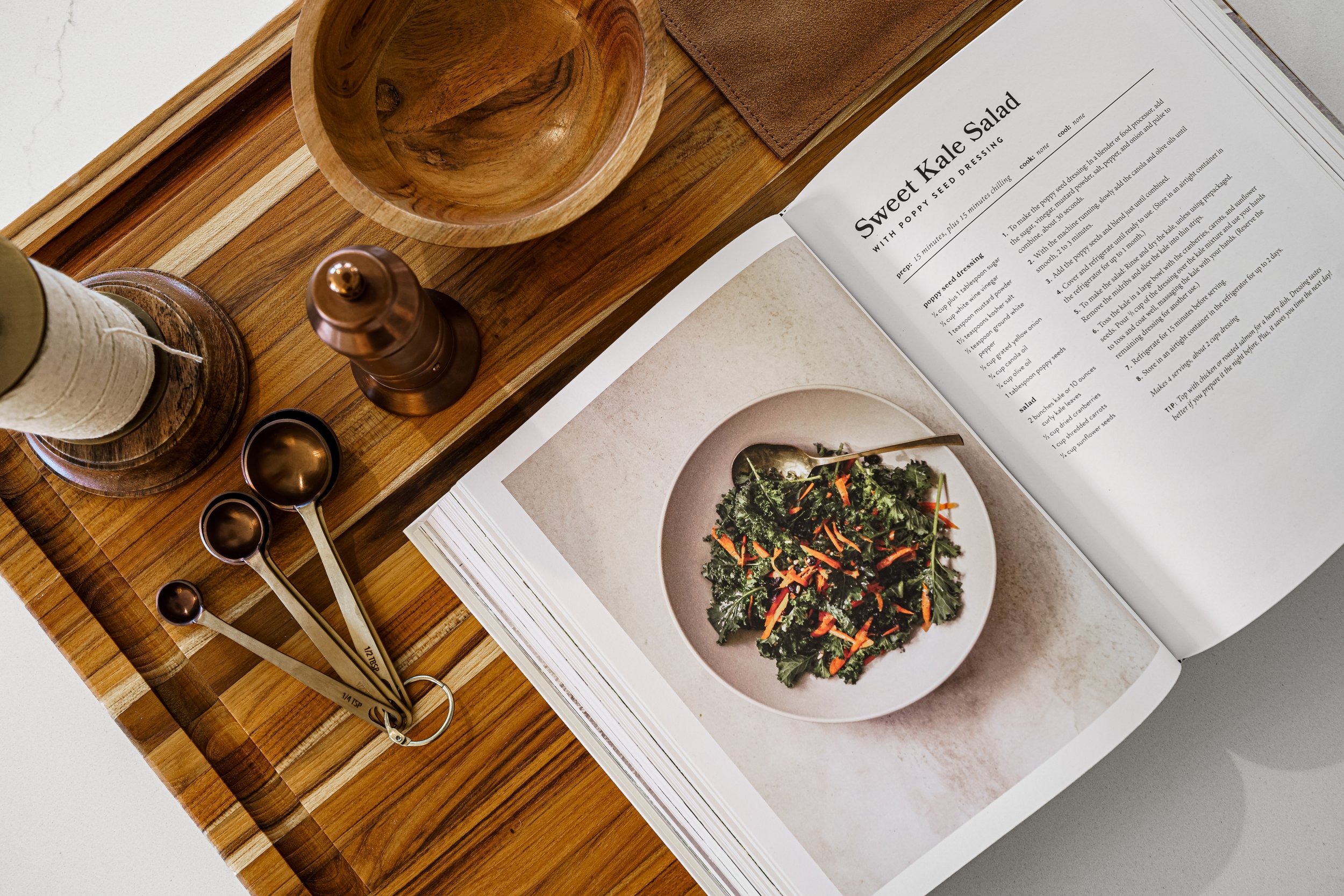 A wooden tray with various kitchen items including a wooden bowl, a small copper container, and a white candle. Next to the tray is an open recipe book with a kale salad recipe, featuring a picture of a kale salad with shredded carrots and other vege