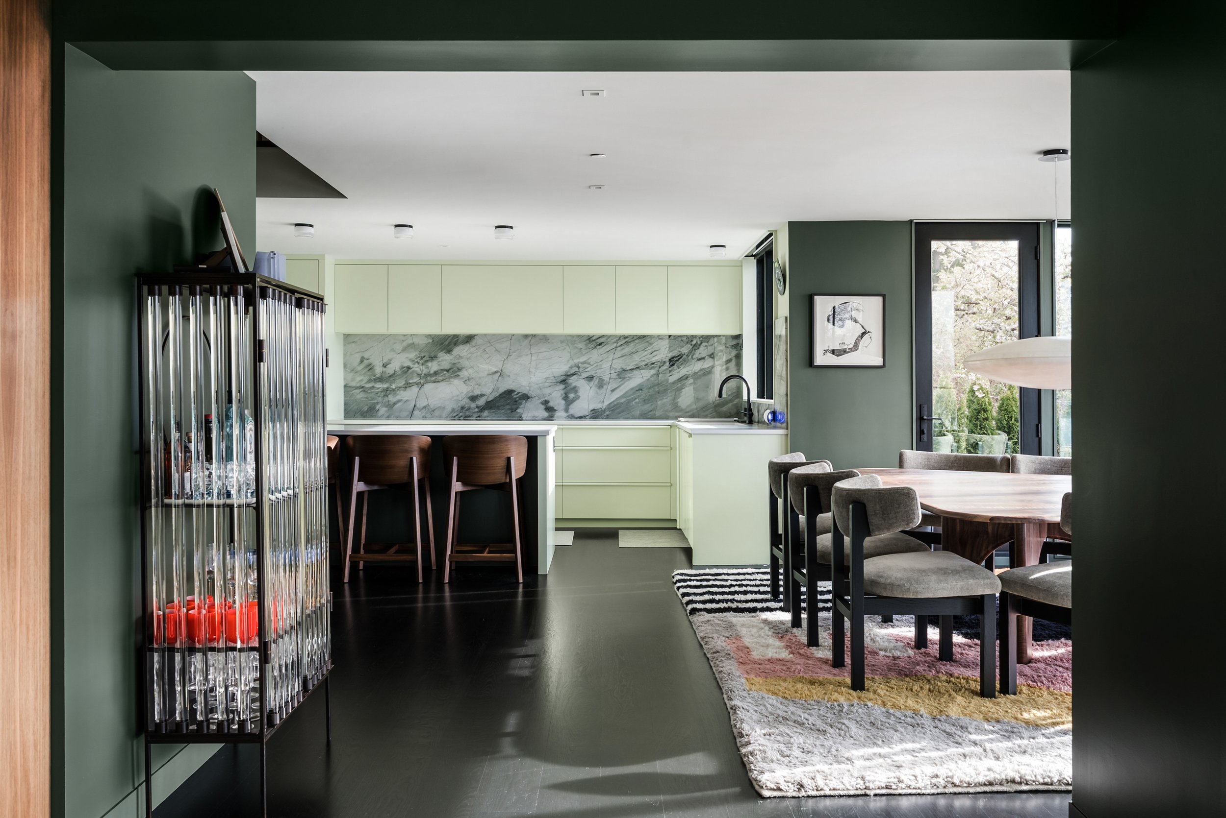 Interior view of a modern kitchen and dining room with green and white walls, dark flooring, a dining table with chairs, and a large window showing outdoor trees.