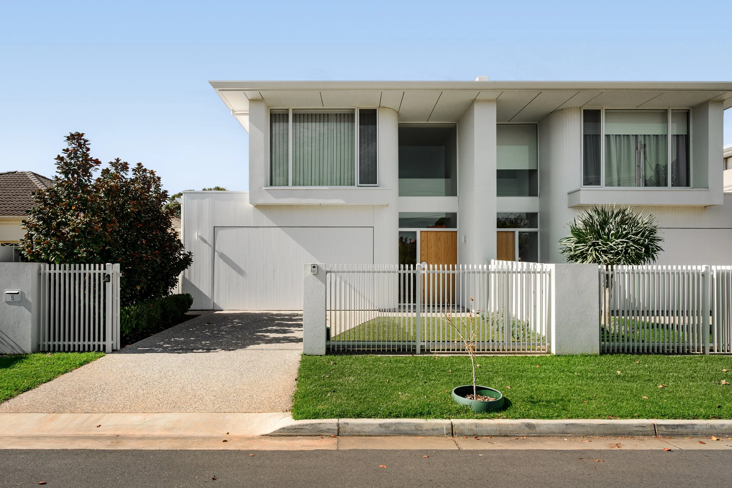 Modern two-story house with a white exterior, large windows, and a white fence. Small front yard with a young tree and well-maintained lawn.