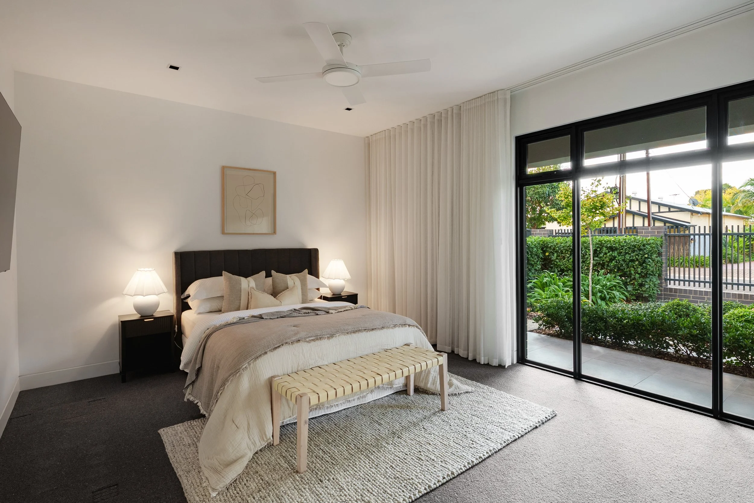A modern bedroom with a bed, nightstands with lamps, a bench at the foot of the bed, a framed abstract line drawing on the wall, large glass sliding doors with white curtains, and outside greenery.
