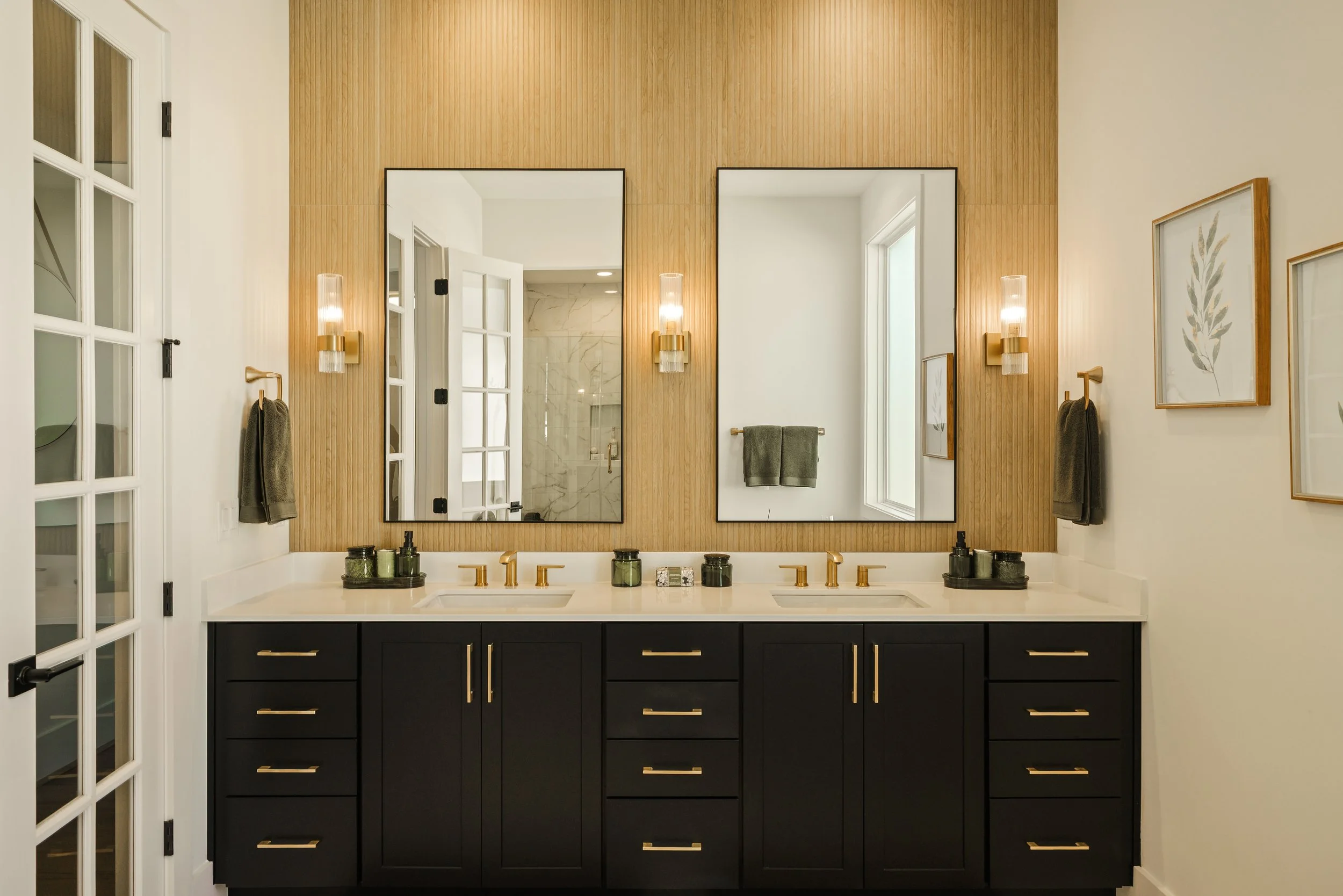 Bathroom vanity with two sinks, black cabinets with gold handles, a white countertop, and large mirrors. The wall behind the mirrors is wood-paneled, and there are wall-mounted lights and framed artwork on the side walls. A towel is hanging on a ring