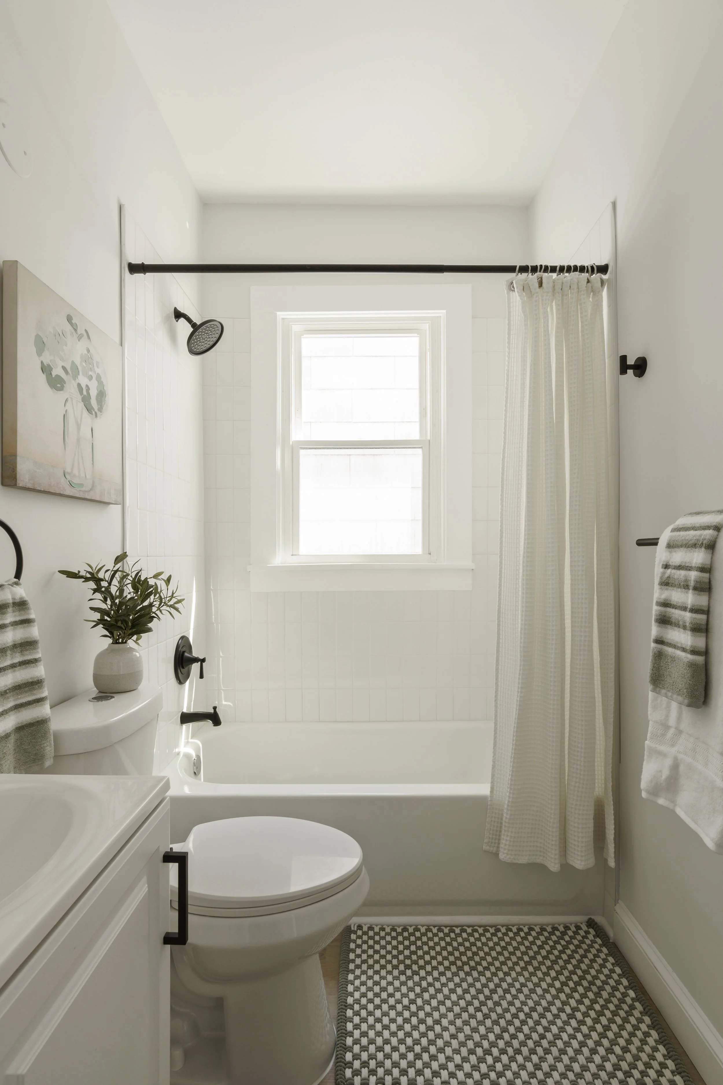 A small, bright bathroom with a white toilet, a white vanity with a cabinet, a potted plant on the toilet tank, a framed artwork on the wall, a window with white trim, a shower with a curtain track, and two towels on the wall hooks.