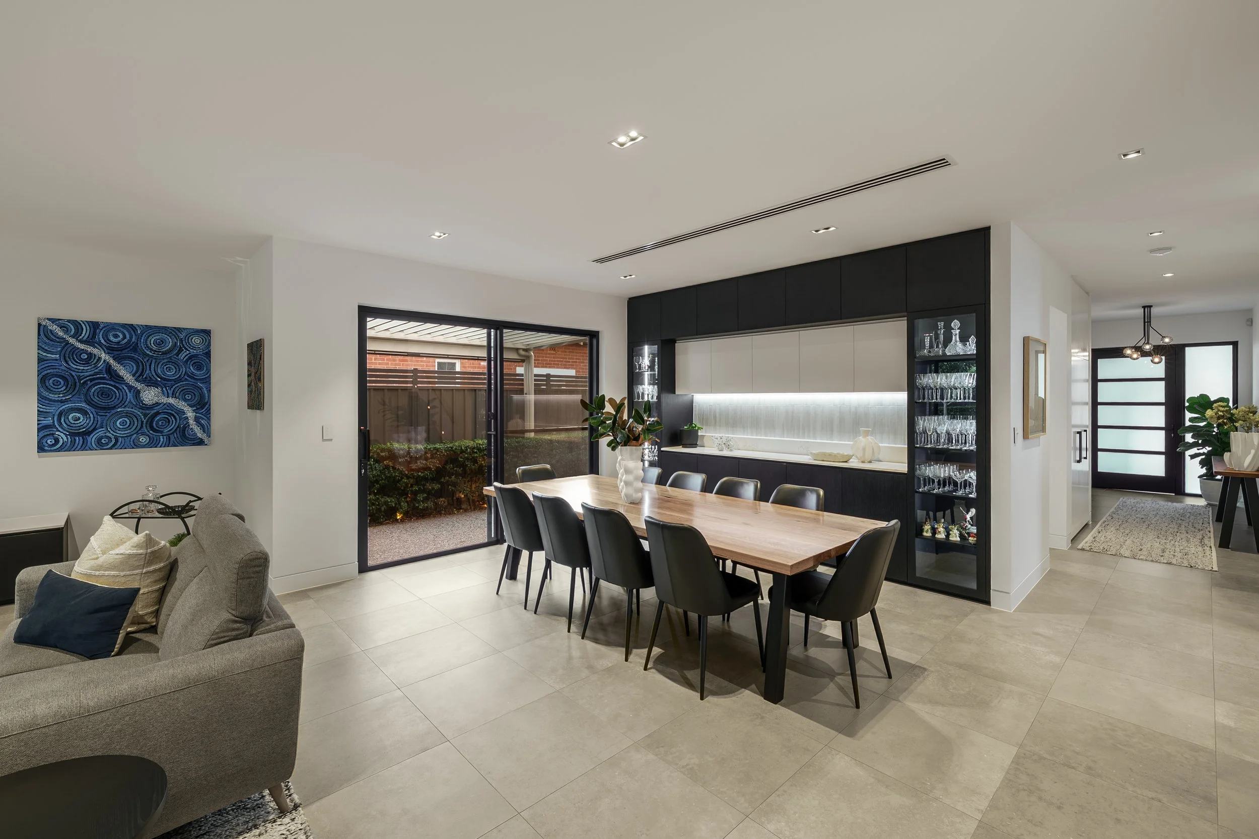 Modern open-plan living and dining area with a wooden dining table, black chairs, a built-in display cabinet with glassware, sliding glass door leading outside, and wall art.