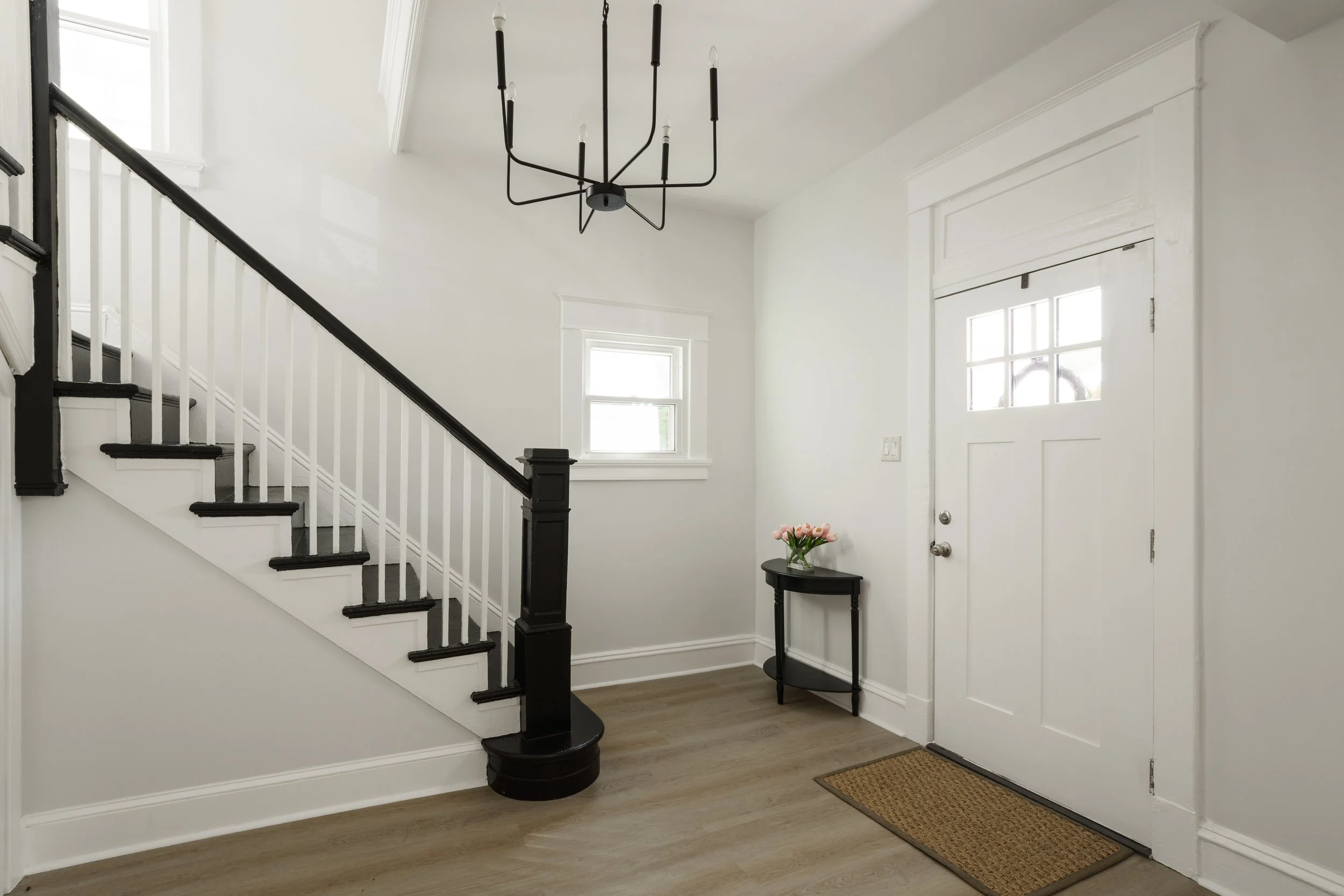 View of a bright entryway with white walls, a white door with glass and wood trim, a black chandelier hanging from the ceiling, a small black table with a vase of pink flowers, and a carpeted welcome mat on a wooden floor.
