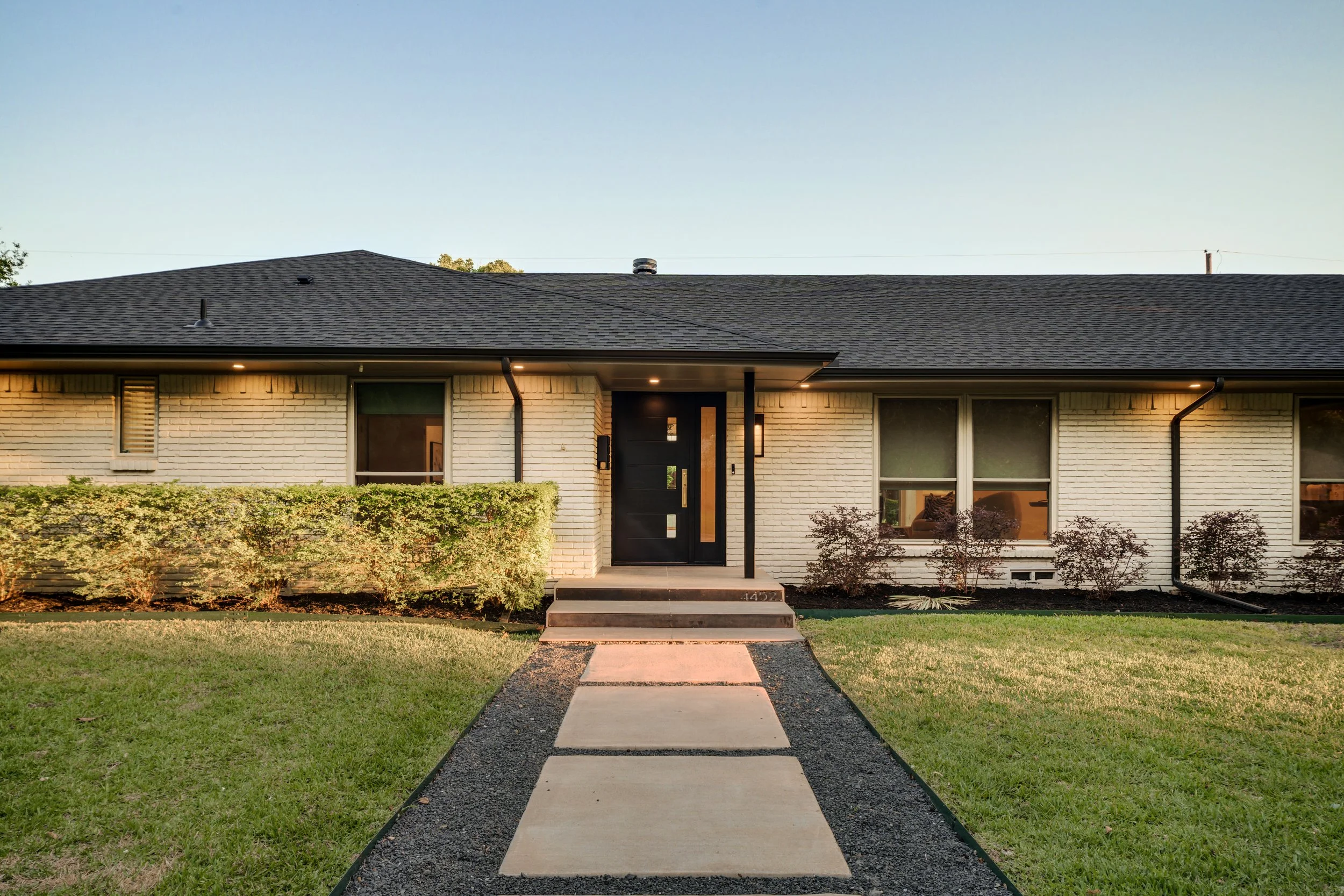 Front view of a modern house with a black door, large windows, a landscaped front yard, and a concrete walkway leading to the entrance during sunset.