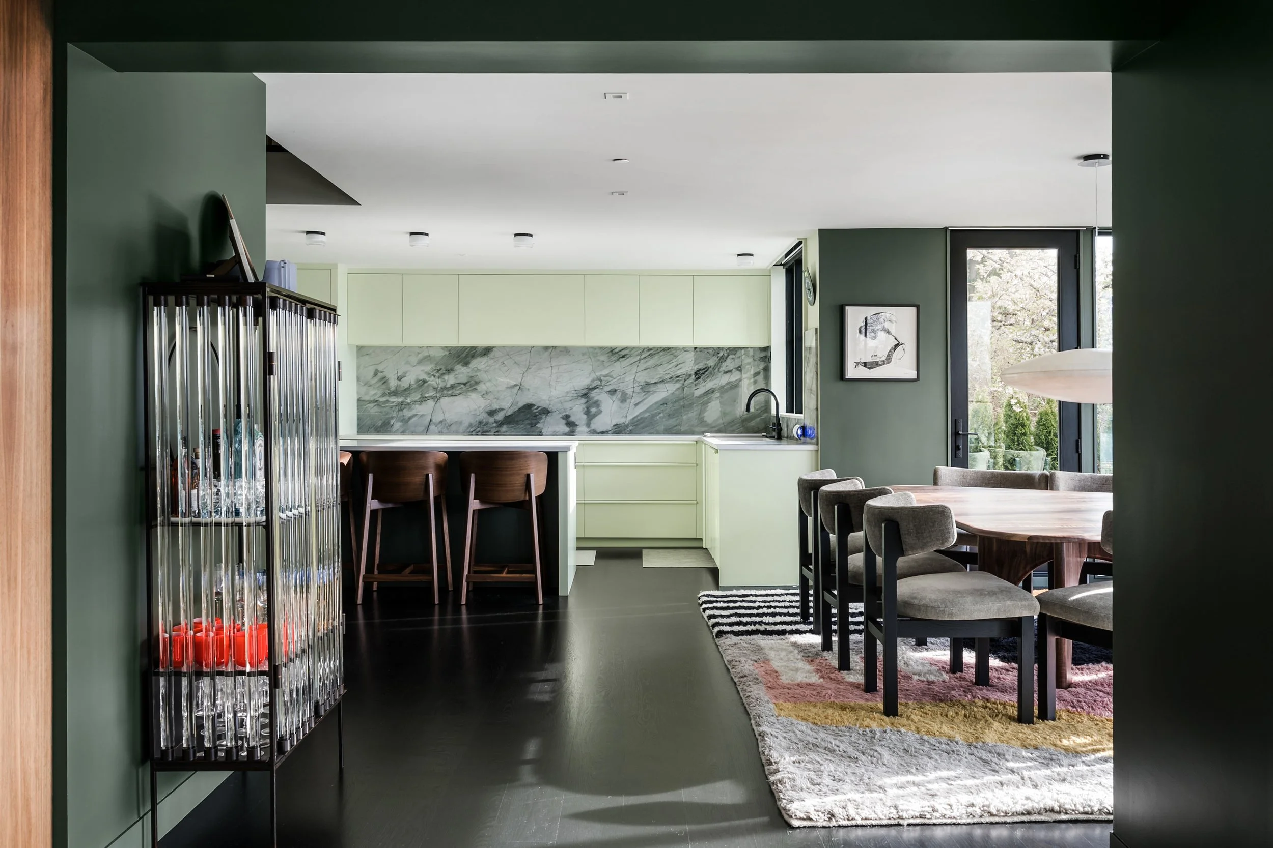 Modern open-concept kitchen and dining area with green and white cabinetry, marble backsplash, black countertops, and a mix of wooden and upholstered chairs. Large windows provide natural light, with a framed black and white art piece on the green wa