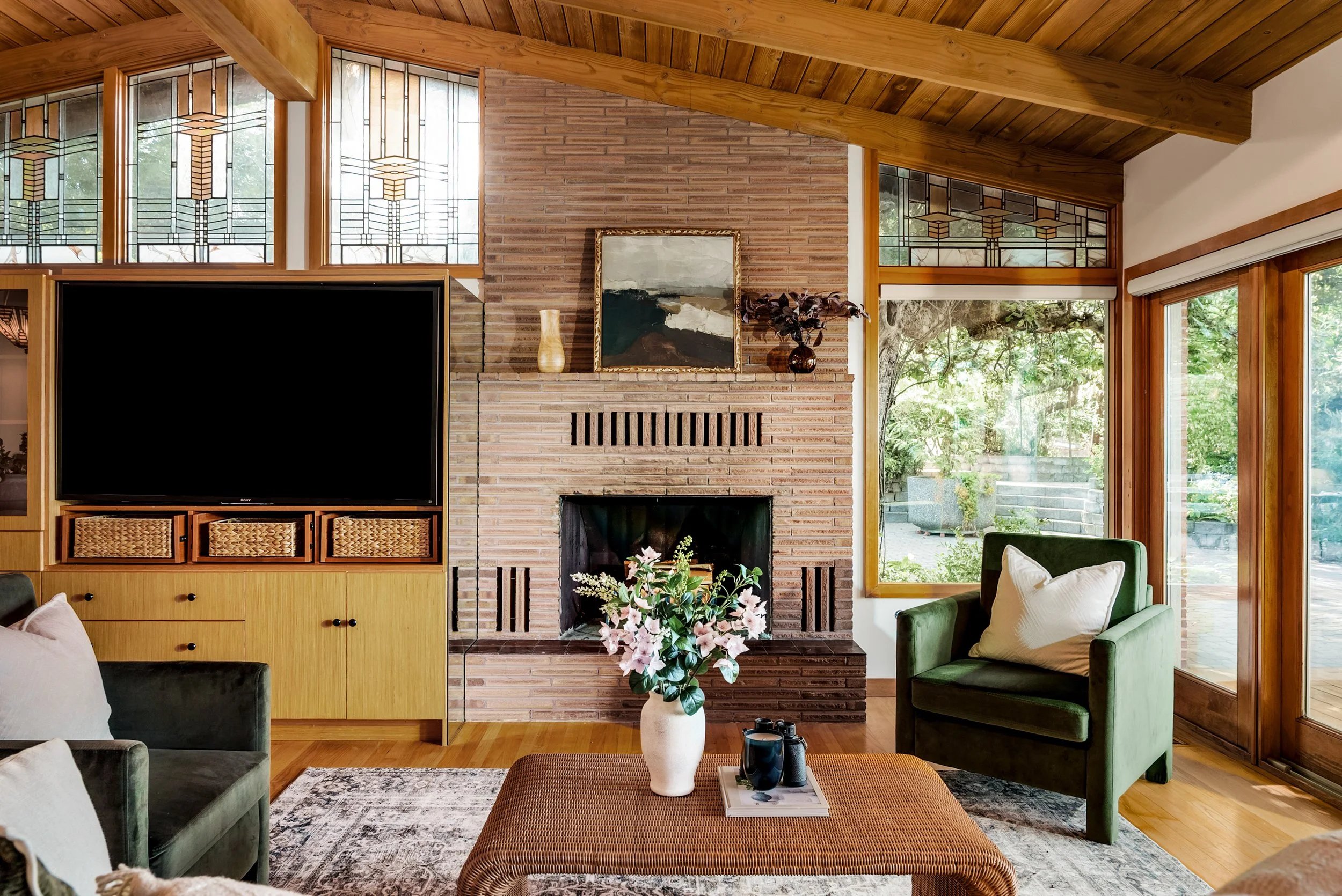 Living room with wooden ceiling, brick fireplace, large windows, green armchairs, a coffee table with a vase of pink flowers, and a view of greenery outside.