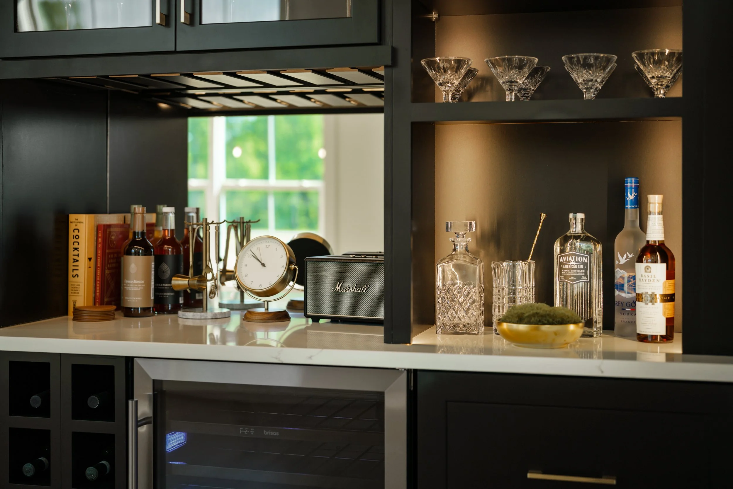 A black kitchen cabinet with glassware, liquor bottles, books, a clock, and a small speaker on a white countertop. A window with green trees outside is visible in the background.