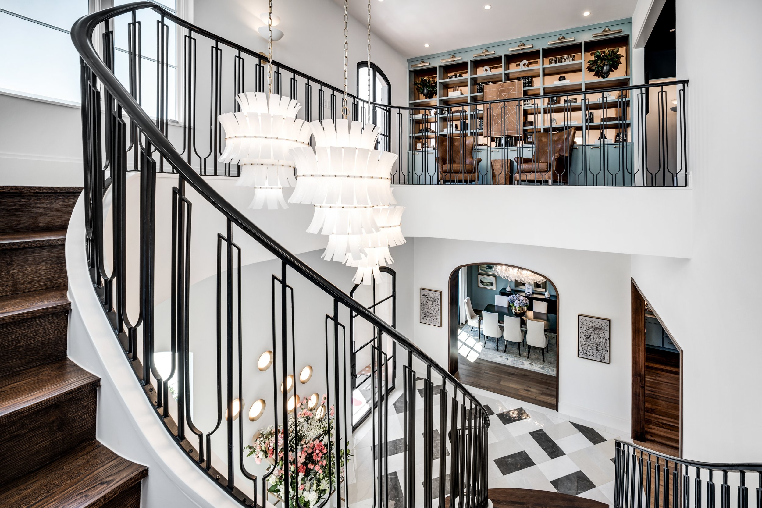 Interior view of a modern home with a curved staircase, black iron railing, white walls, and a chandelier. The area features a living space with top shelves, two chairs, and a small table. There is a dining area with white chairs visible below.
