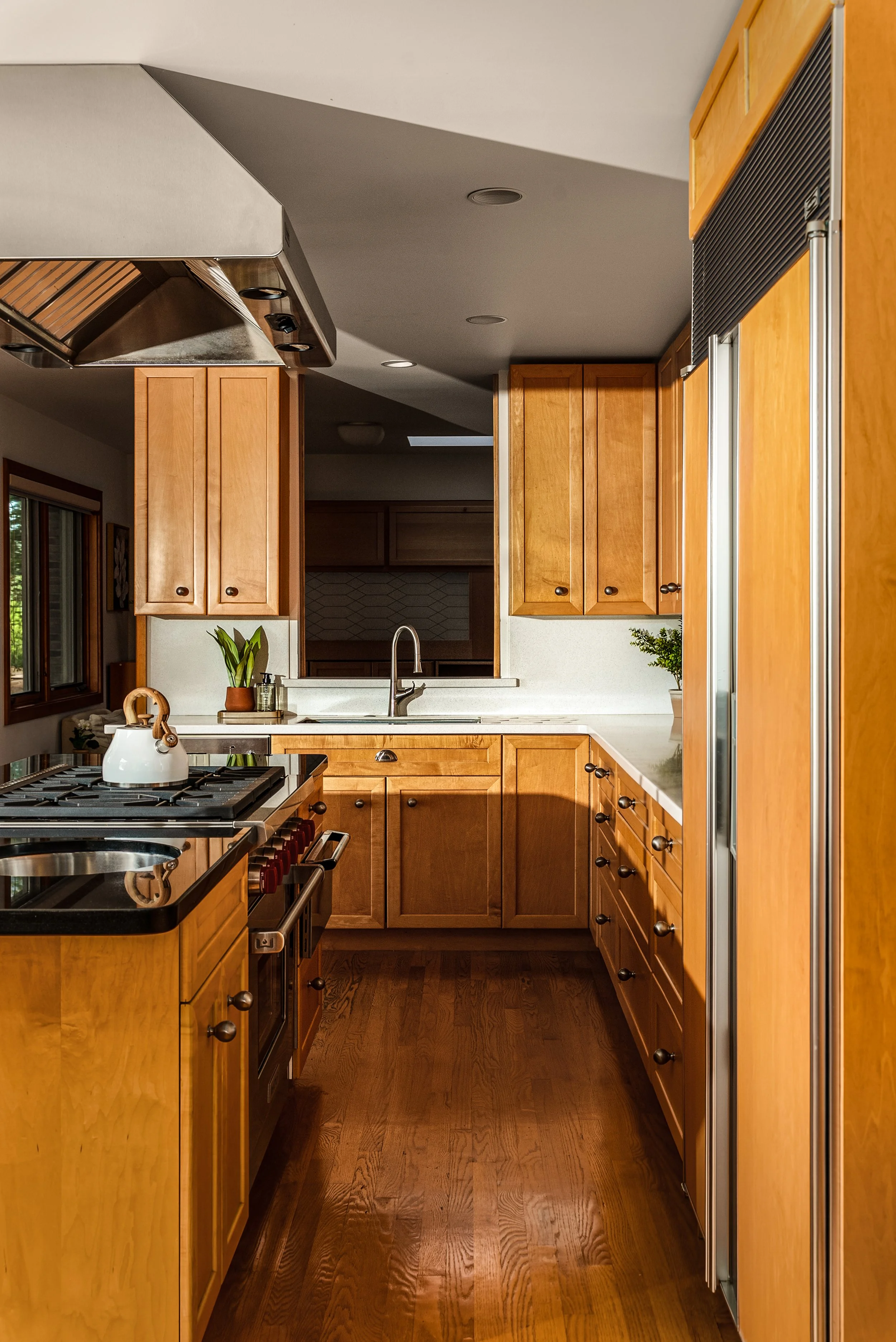A kitchen with wooden cabinets, a stove with a teapot, a sink area with plants, and hardwood flooring.
