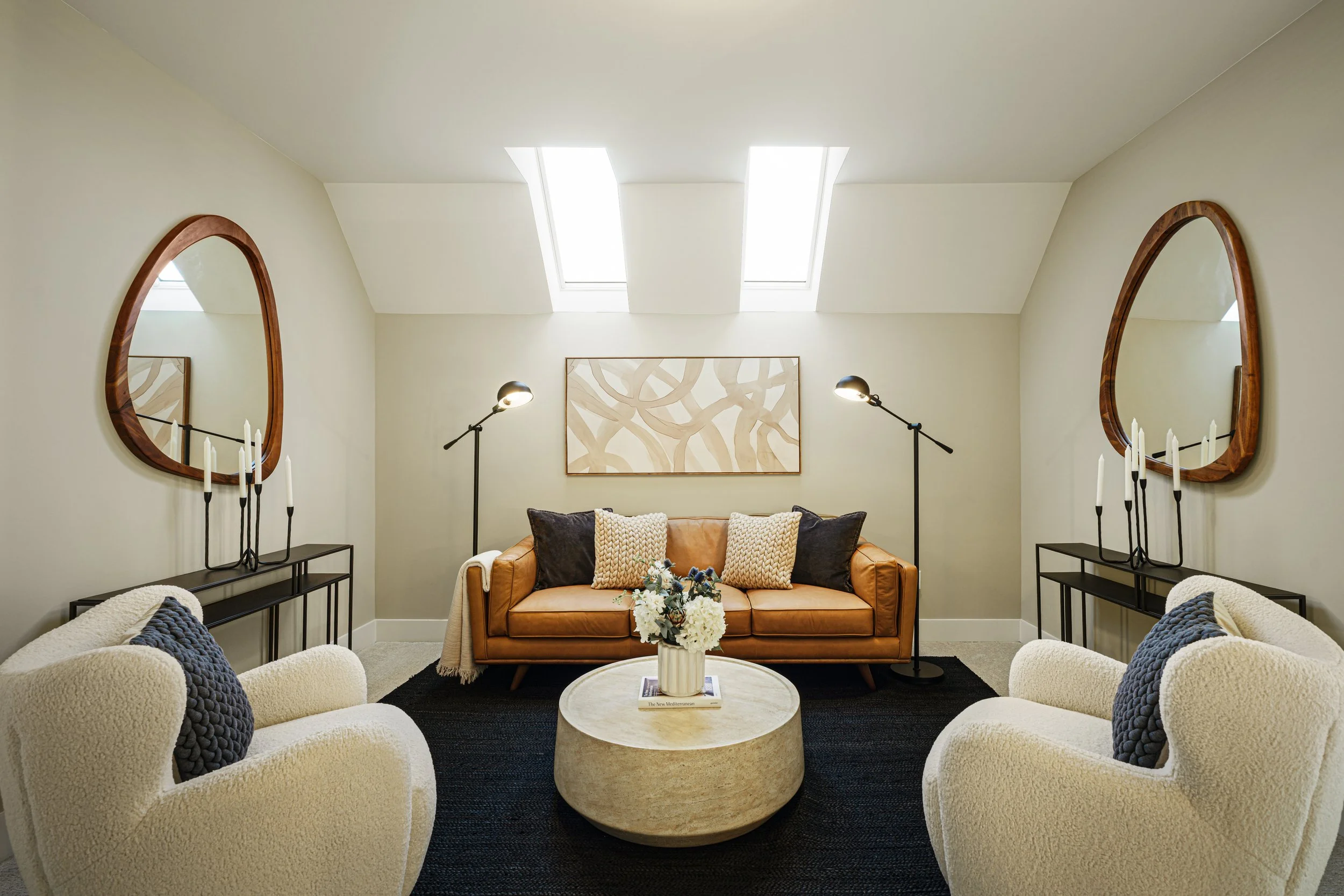 A cozy living room with white armchairs, a tan leather sofa, black side tables, modern table lamps, a wooden abstract wall art, and two oval-shaped mirrors with wooden frames, illuminated by two skylights.