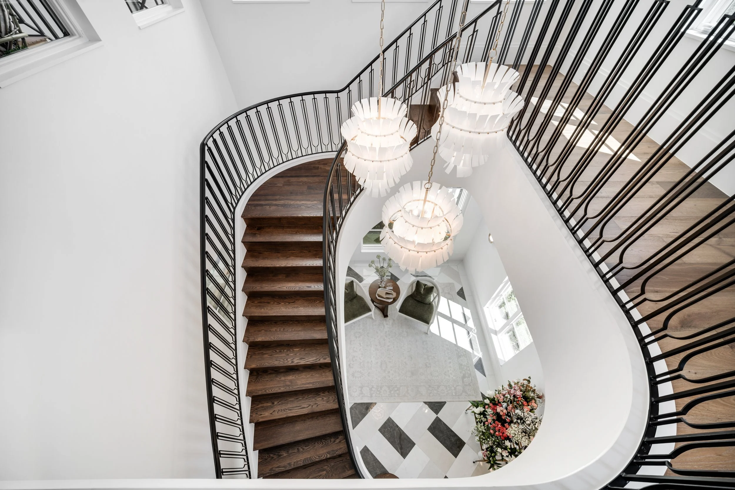 Interior view of a multi-story foyer showing a staircase with dark wood steps and black metal railing, a large modern chandelier hanging from the ceiling, a sitting area with armchairs and a table, large windows allowing natural light, and elegant de