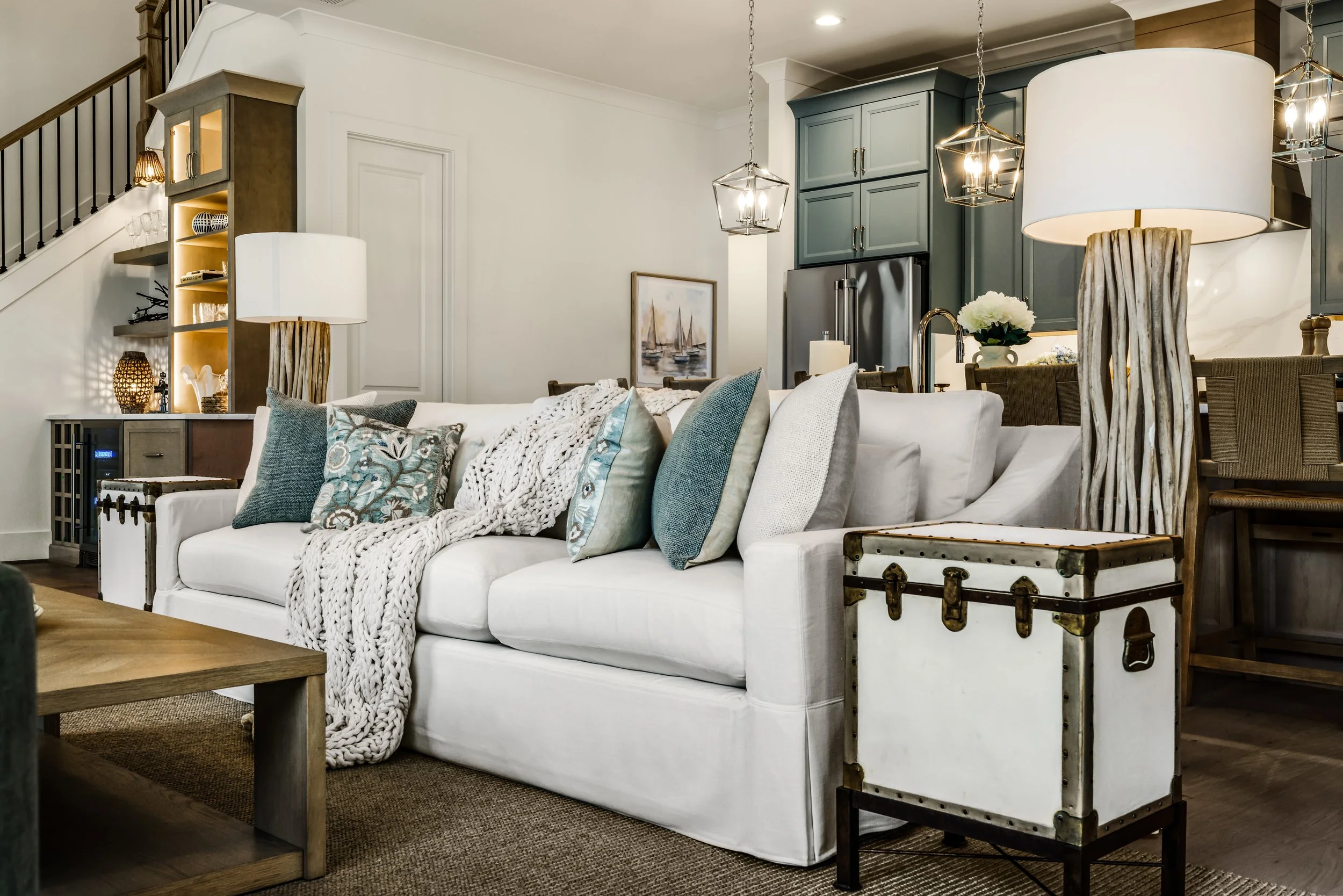 Living room with a white sofa, decorative pillows, a wooden coffee table, two large wicker lamps, a paint painting of sailboats on the wall, a built-in bookshelf, and a kitchen visible in the background with blue cabinets and stainless steel refriger