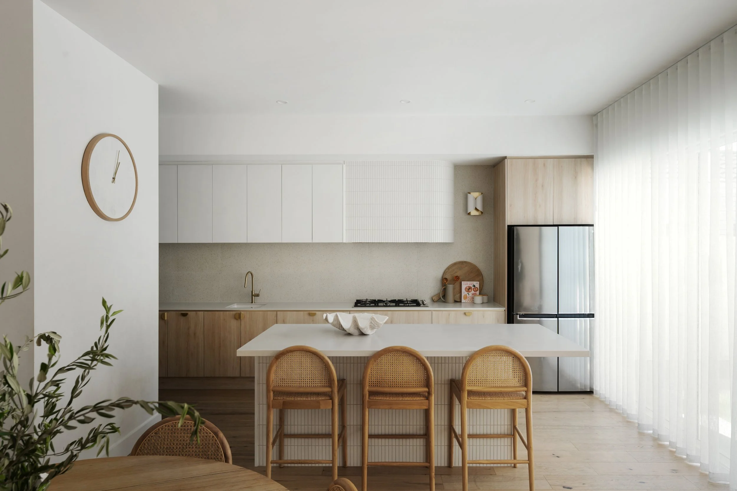 Modern kitchen with white cabinets, wooden accents, and a stainless steel refrigerator. A round wall clock and a dining table with woven chairs are visible, with sheer curtains on large windows.