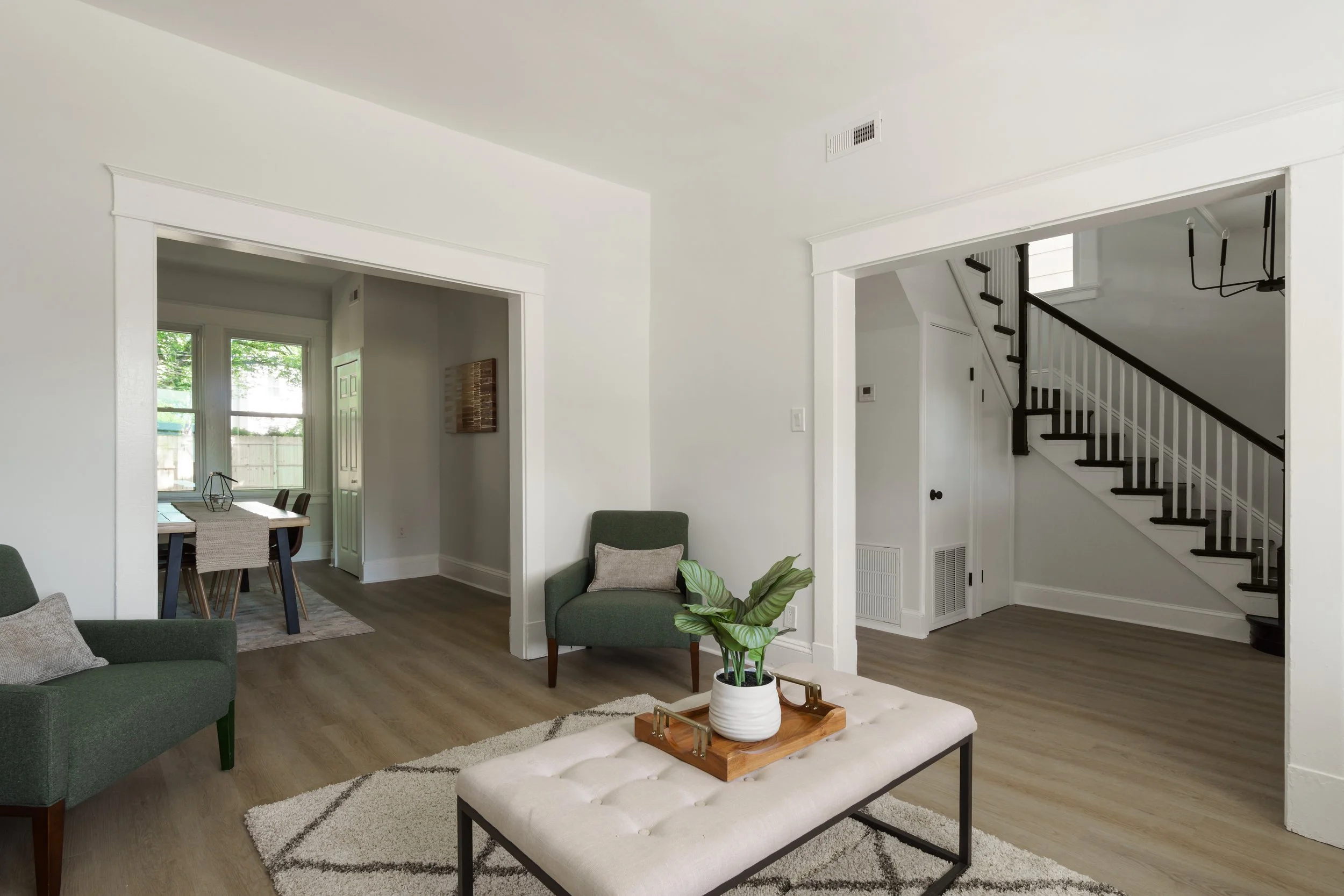 Living room with green armchair, white tufted ottoman with tray and plant, hardwood floors, and a staircase leading upstairs.