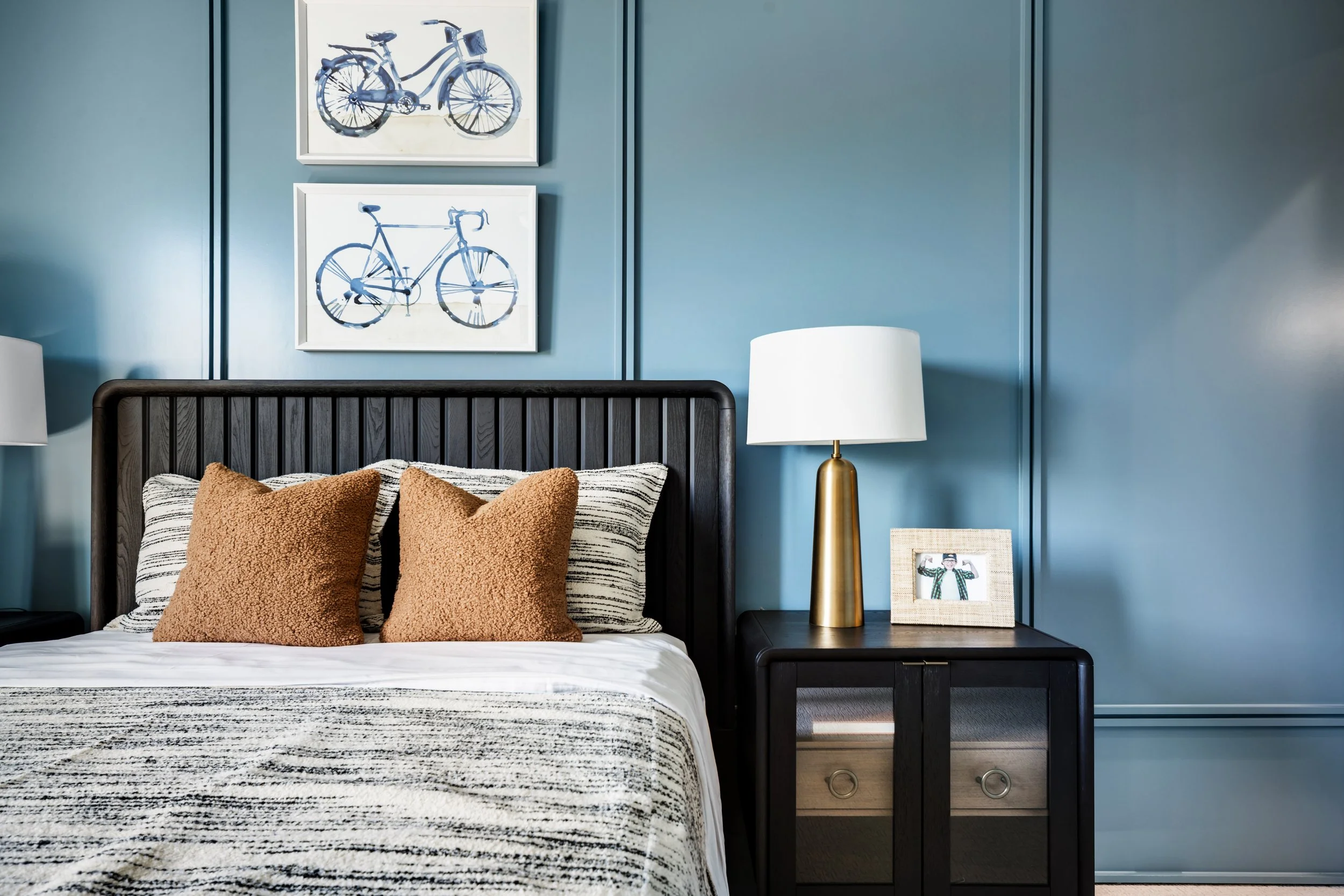 A bedroom with a black bed frame, striped blanket, and four brown pillows. Two framed bicycle artworks are on a blue wall above the bed. A cylindrical gold table lamp and a framed photo of a woman are on a black nightstand beside the bed.