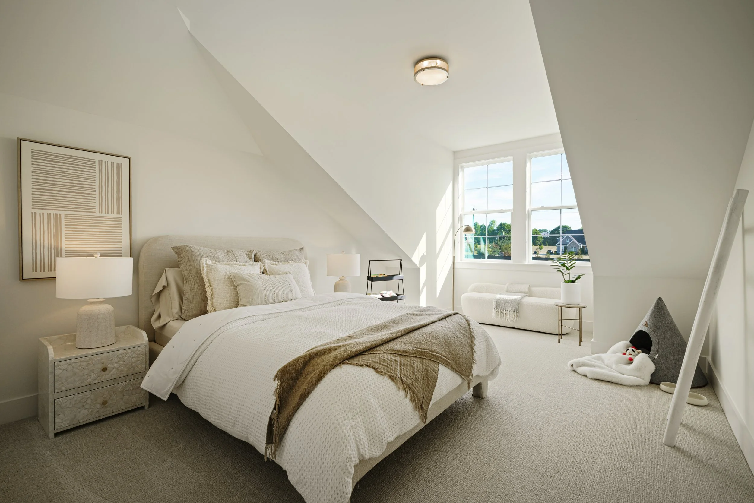 A bedroom with a sloped ceiling, white bed with beige and cream bedding, two nightstands with lamps, a window with a view of a suburban neighborhood, and decorative wall art.
