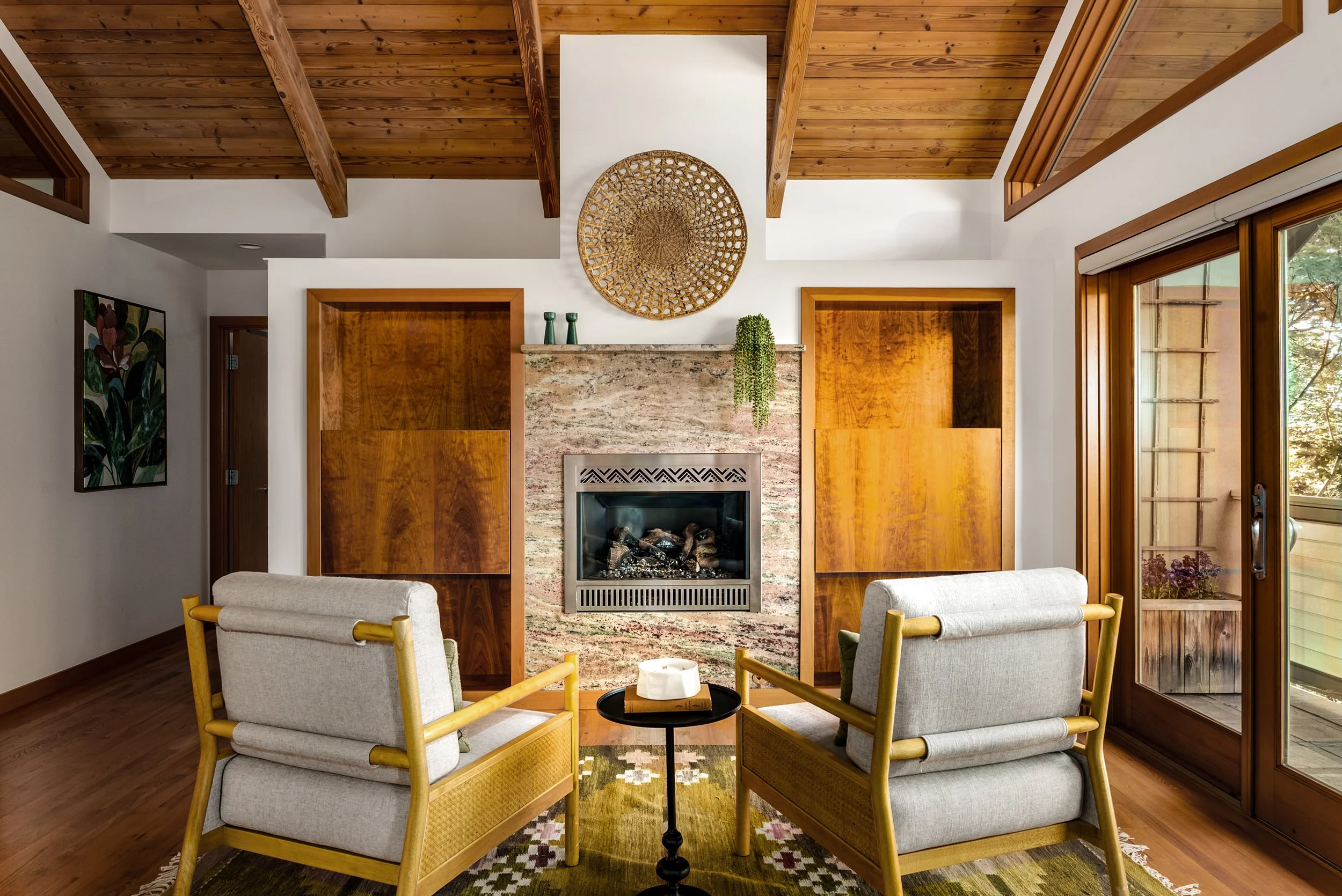 Living room with two beige armchairs around a small black side table, a fireplace with a stone surround at the center, and a decorative circular wall art above it. The room features a wooden ceiling with exposed beams, wood trim around the doors and 