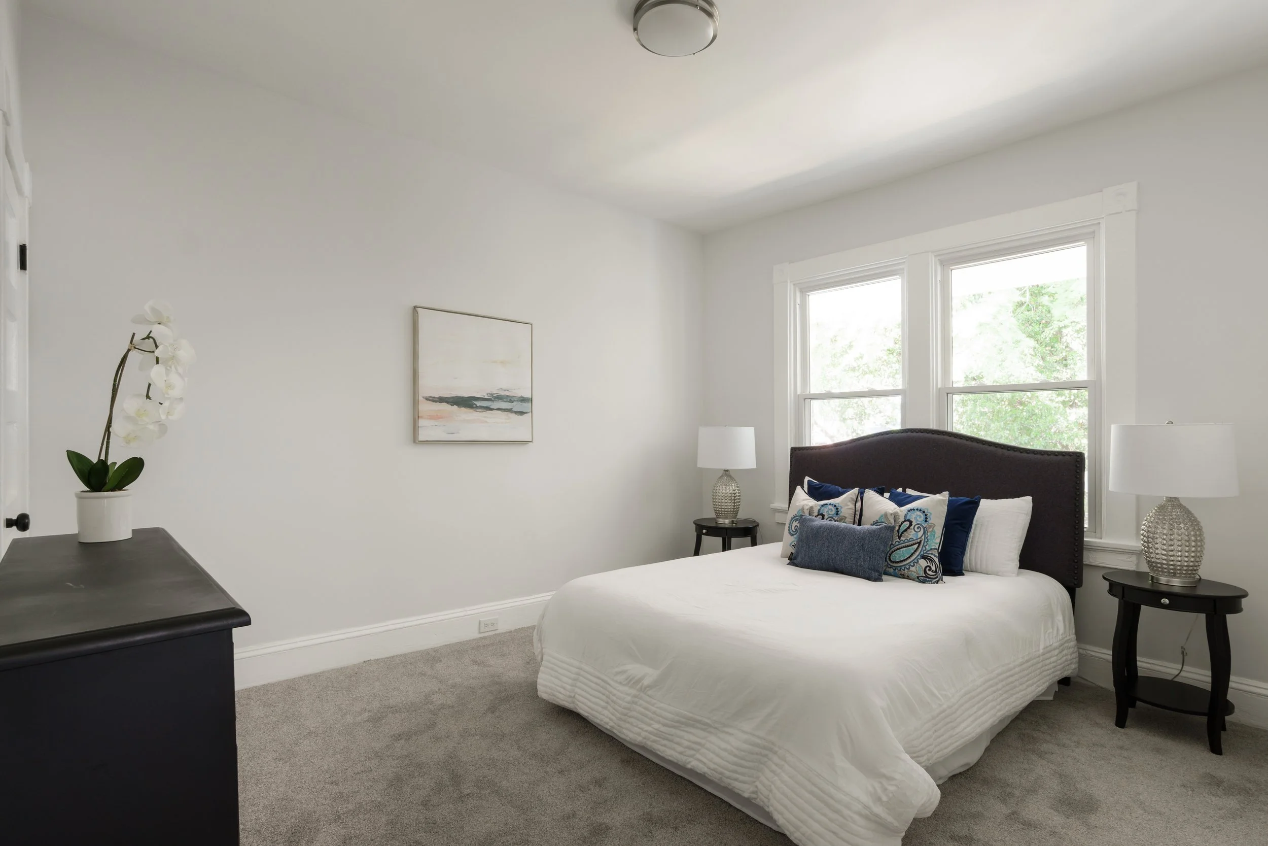 A bright bedroom with a large bed featuring a dark headboard, white bedding, and decorative blue and paisley pillows. Two round black nightstands with matching textured lamps sit beside the bed. There are two large windows with white trim, allowing n