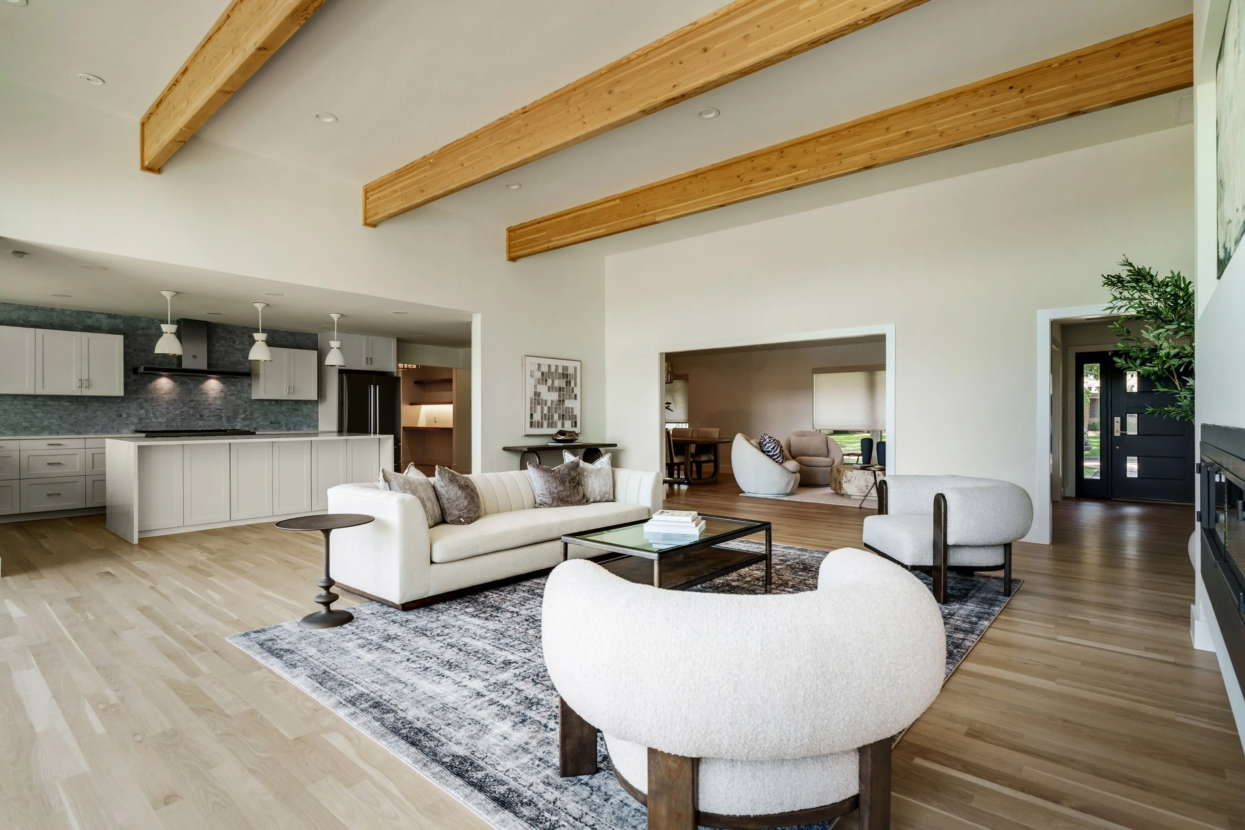 Spacious living room with white sofas, armchairs, wooden beams on the ceiling, and a kitchen area with white cabinetry and pendant lighting.
