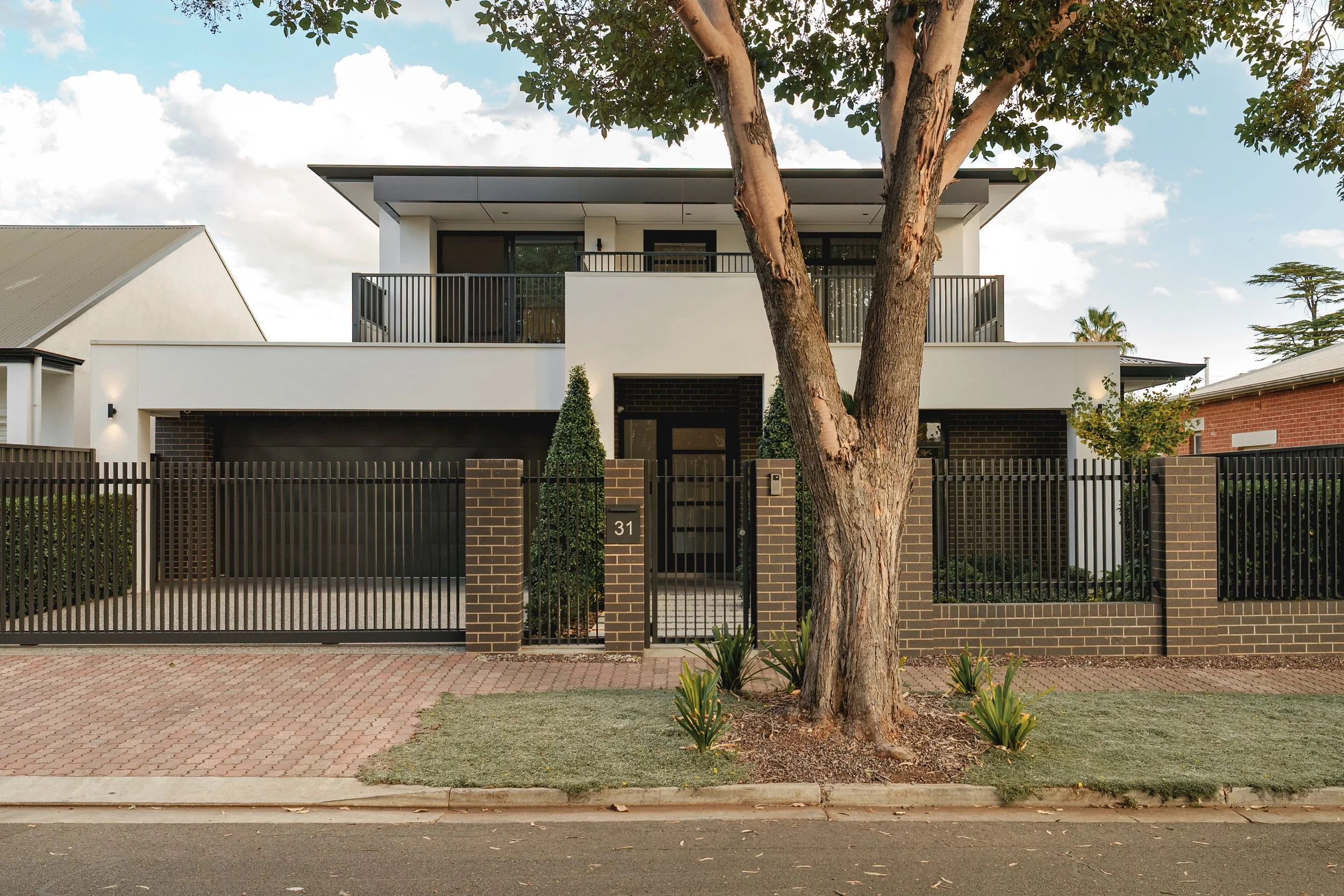 Modern two-story house with white and black exterior, balcony, and black metal fence. Front yard has small plants and trees, with a brick sidewalk and road in front.