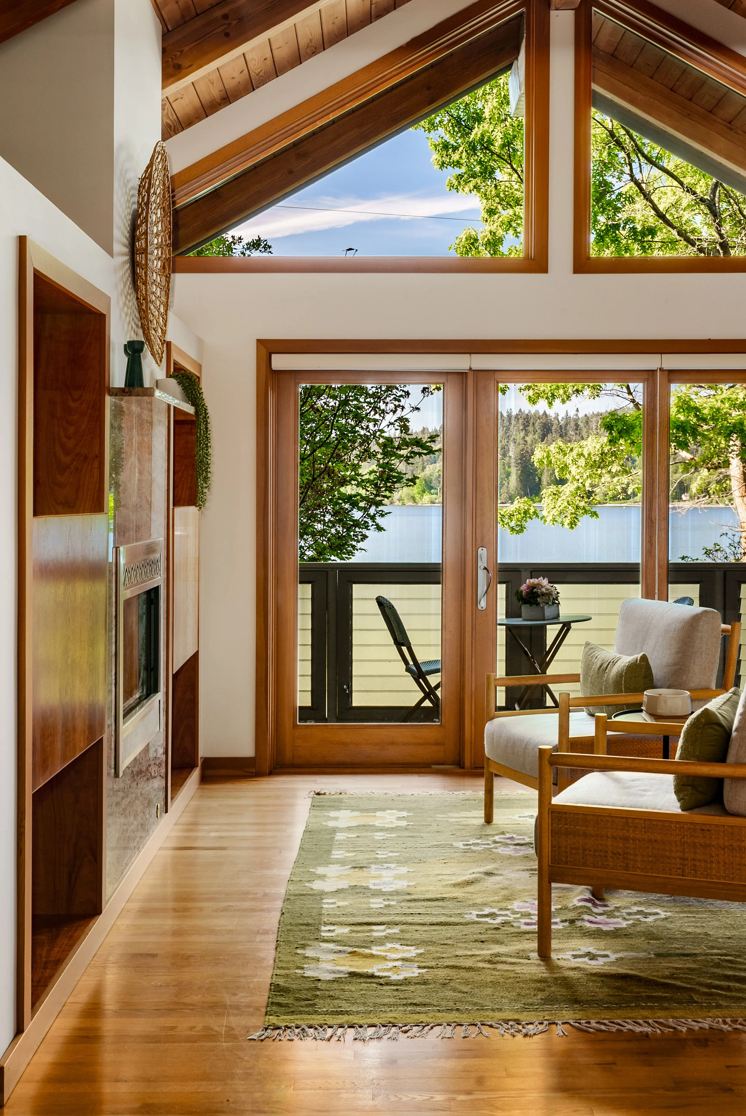 Living room with large windows and sliding door overlooking a lake and trees, wooden ceiling and trim, beige sofa with green and beige pillows, green patterned rug, wooden side table with flowers, and a balcony with chairs.