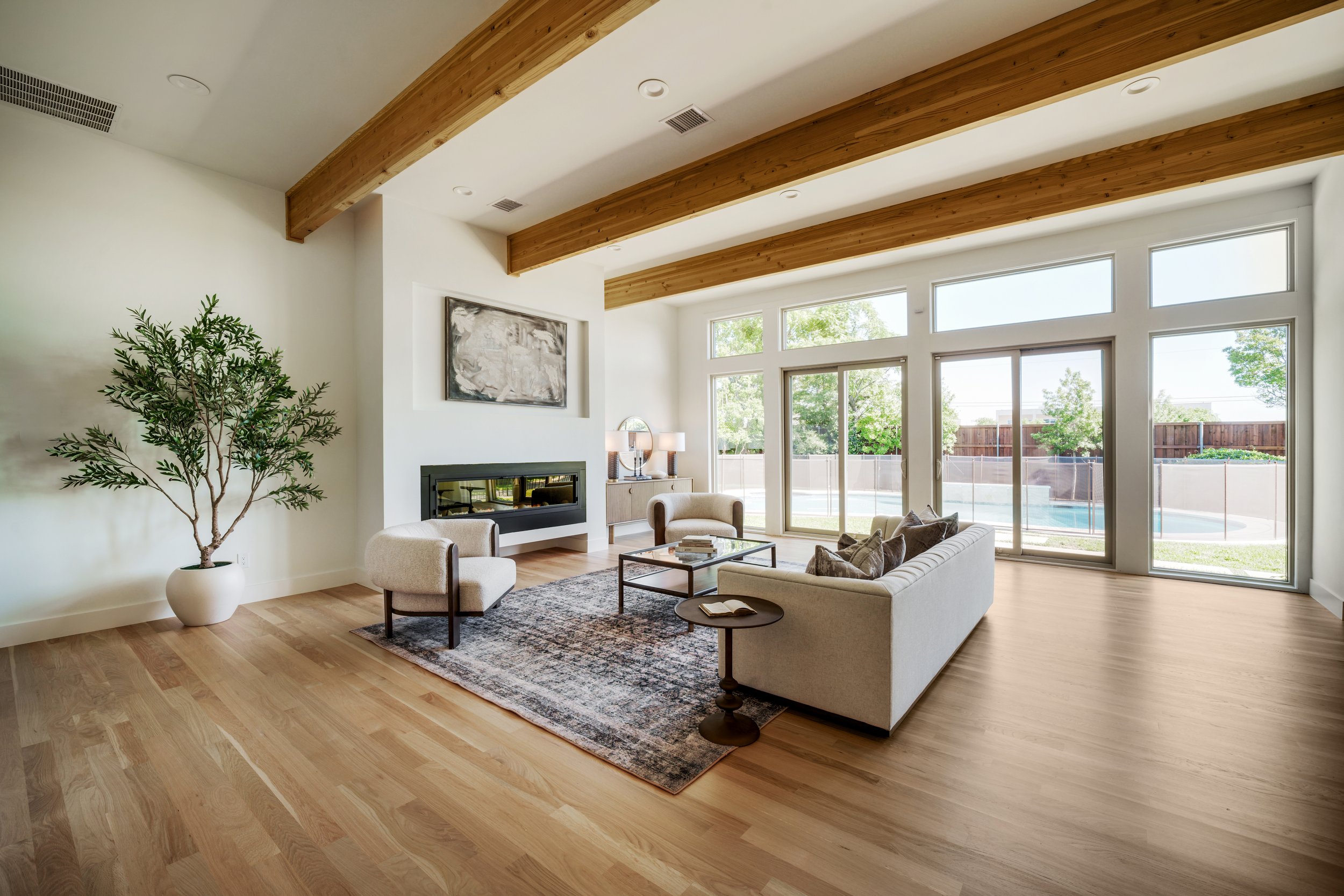 Modern living room with wood beam ceiling, large windows, beige sofa, armchair, fireplace, and decorative plants.