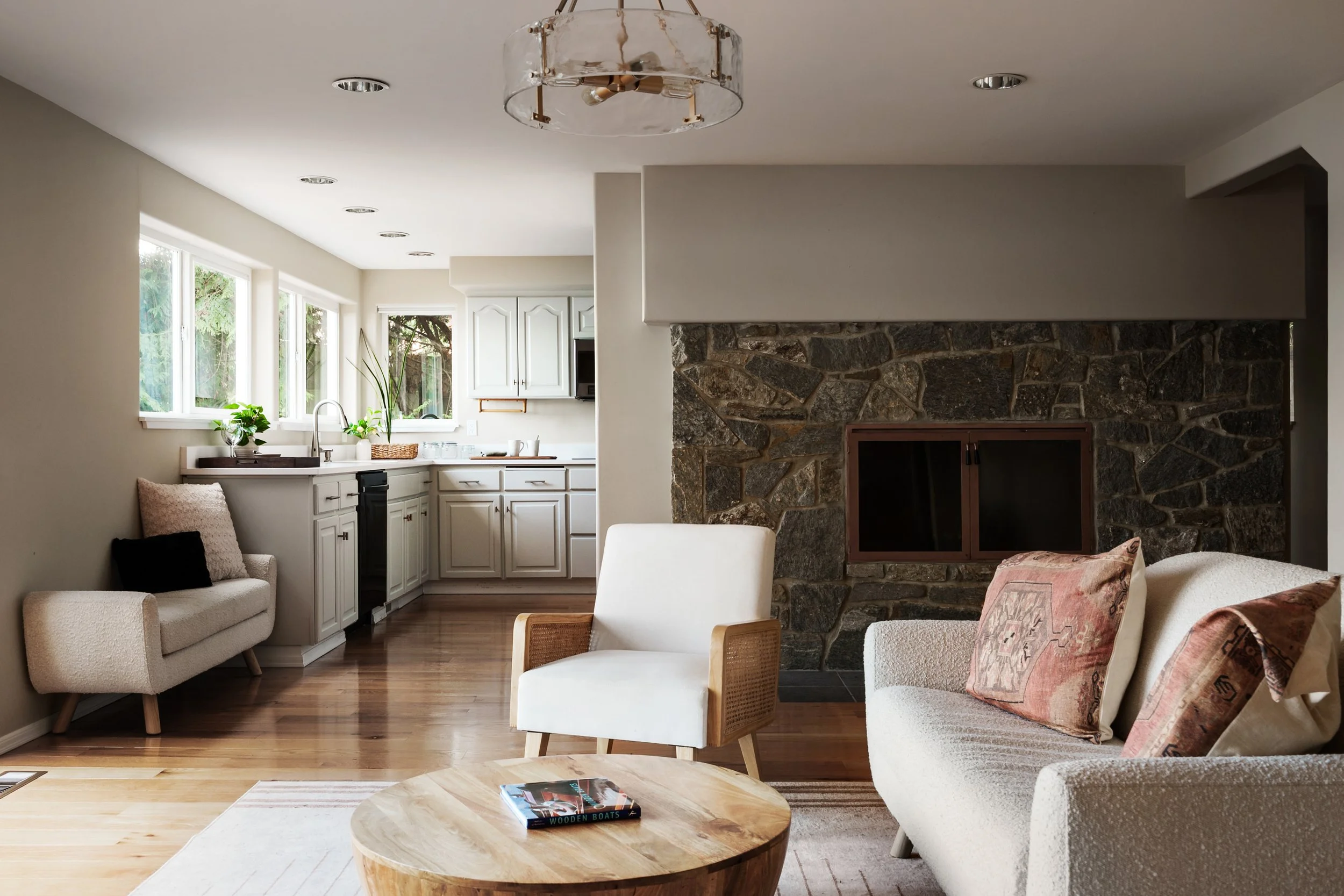 Living room with beige sofas, coffee table with book, fireplace with stone wall, white kitchen cabinets, and large windows with greenery outside.