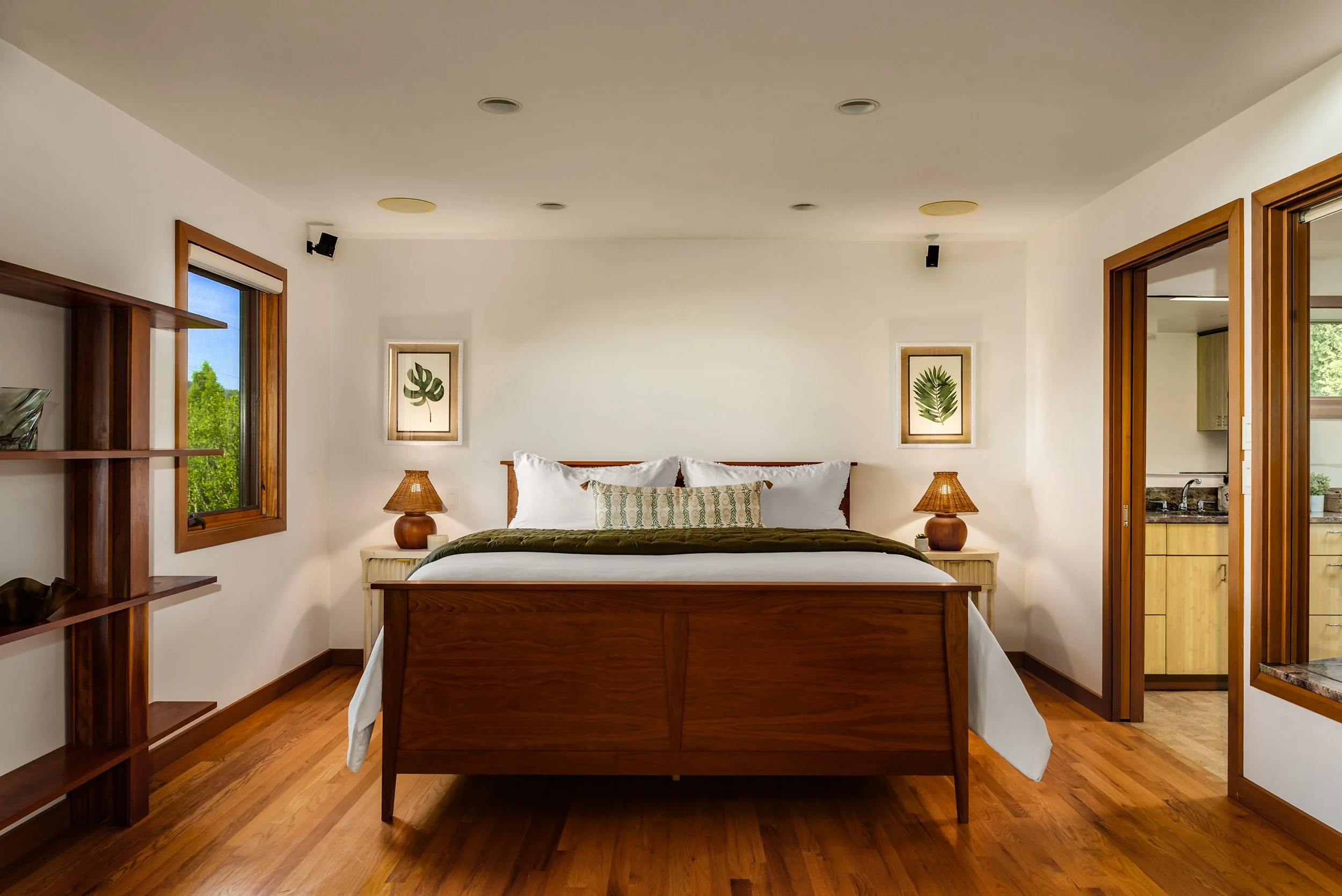 Cozy bedroom with a wooden bed, white bedding, and side tables with lamps. Two framed leaf prints on the wall, windows with wooden frames showing greenery outside, and a doorway leading to a kitchenette.