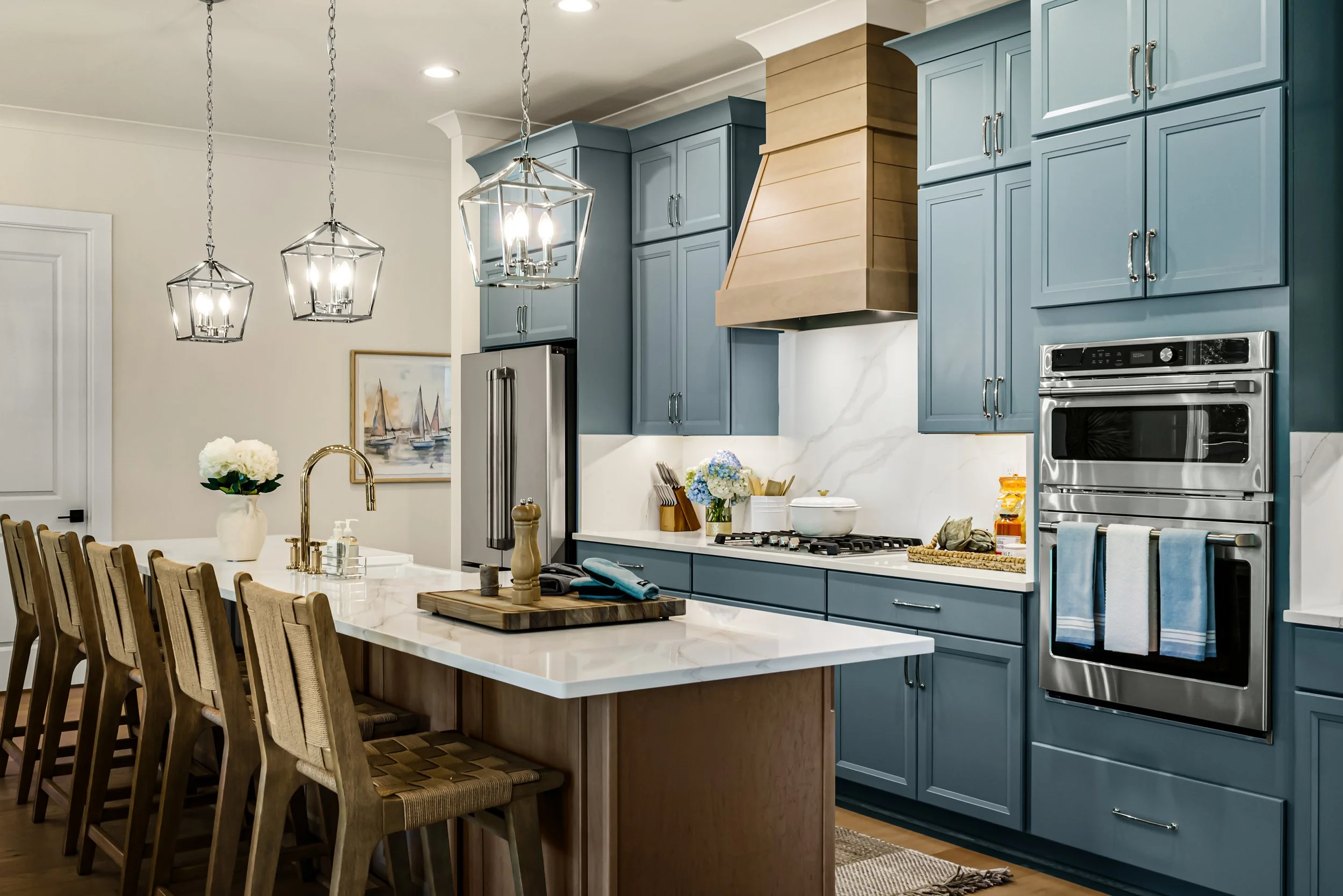 Modern kitchen with blue cabinets, stainless steel appliances, a white marble countertop, and two geometric pendant lights hanging above a dining table with wooden chairs.