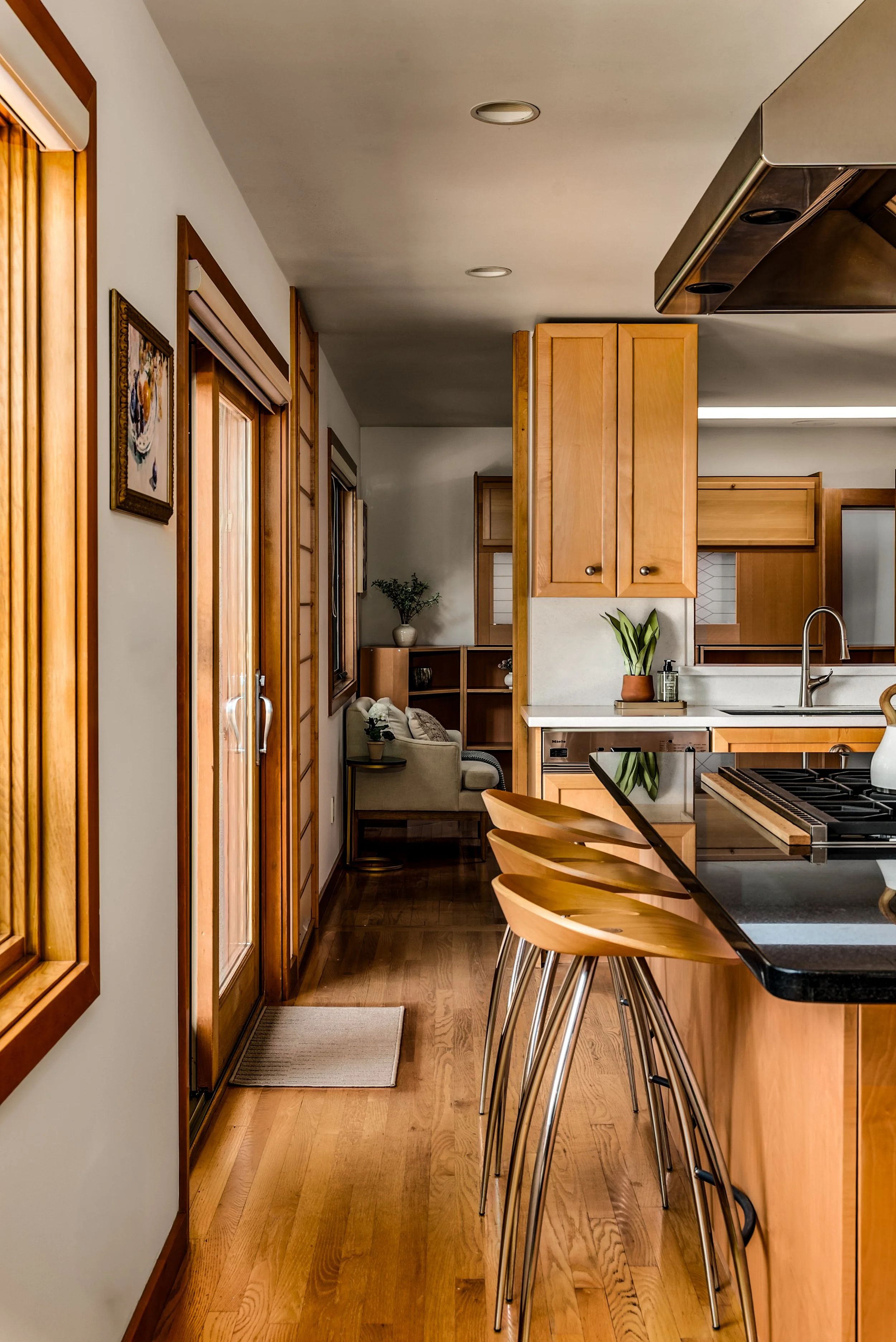 Interior of a kitchen and dining area with wooden cabinets, a black countertop with barstools, and sliding glass doors with wooden frames.
