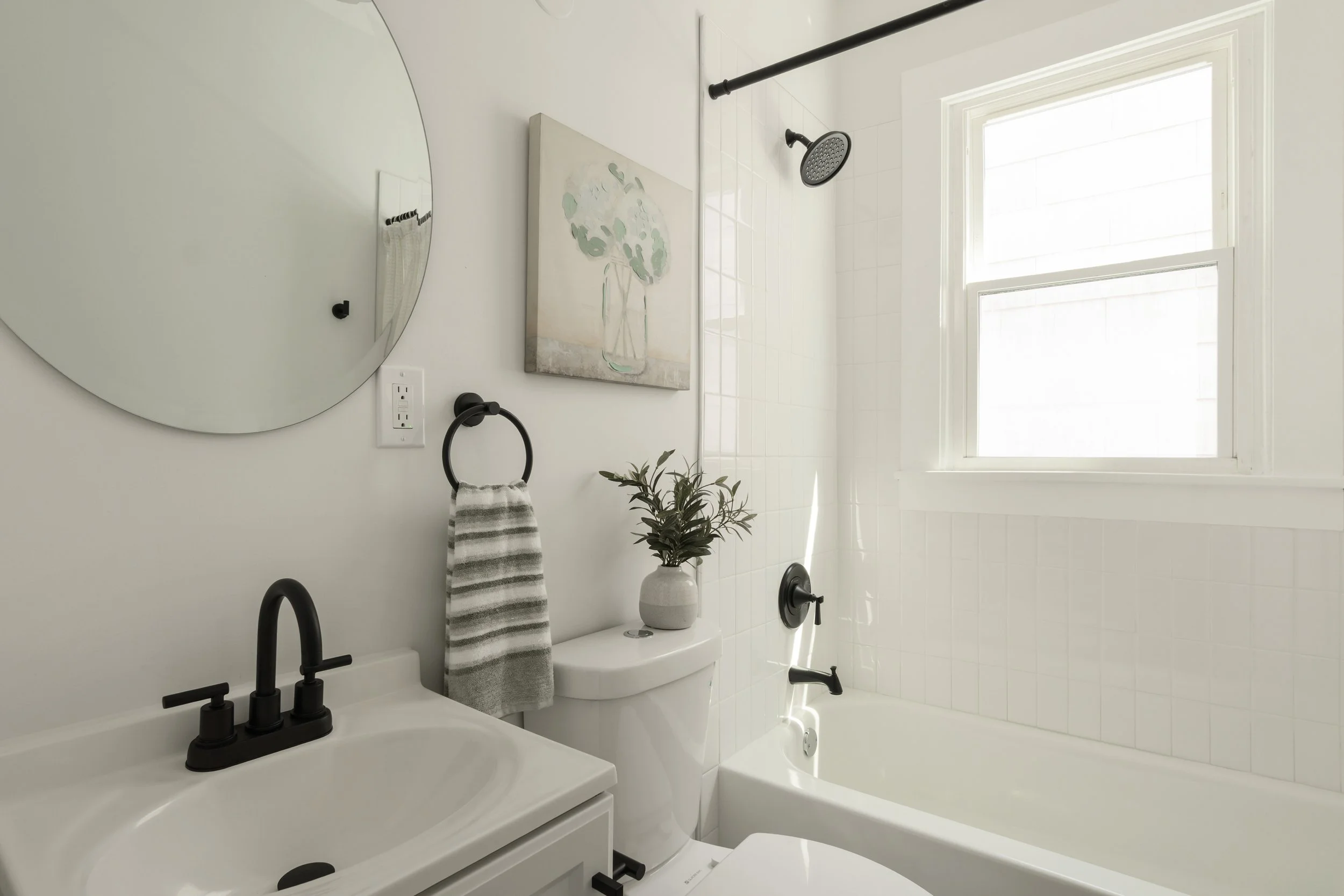 Bright bathroom with white tiles, a bathtub, a window, a round mirror, a black faucet, a towel ring with a striped towel, a painting of a vase with greenery, a potted plant, and modern fixtures.
