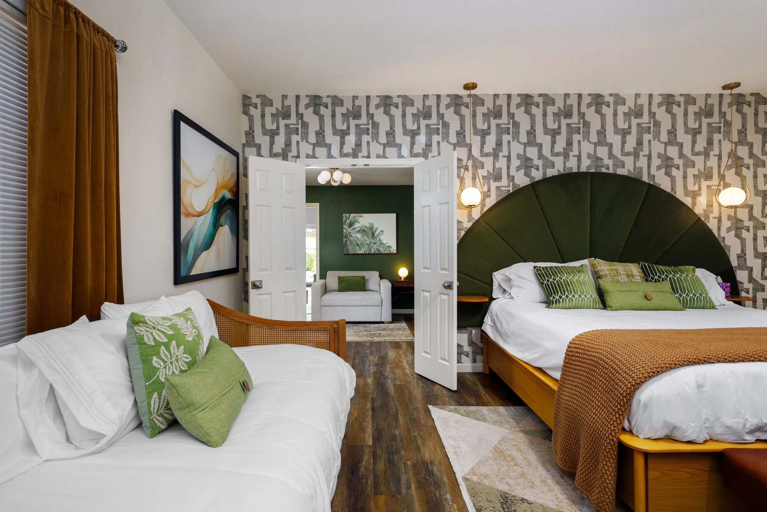 A cozy bedroom with two beds, green accent pillows, a large green upholstered headboard, and modern lighting. There is a view into an adjacent room with a white couch, green pillow, and artwork of palm trees on the wall.
