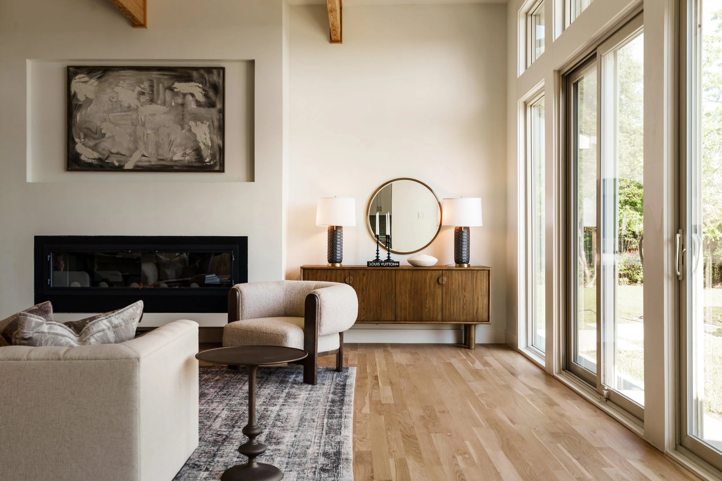 Living room with beige sofa, round side table, area rug, wall art, side table with lamps, round mirror, and large sliding glass doors showing a green outdoor scene.