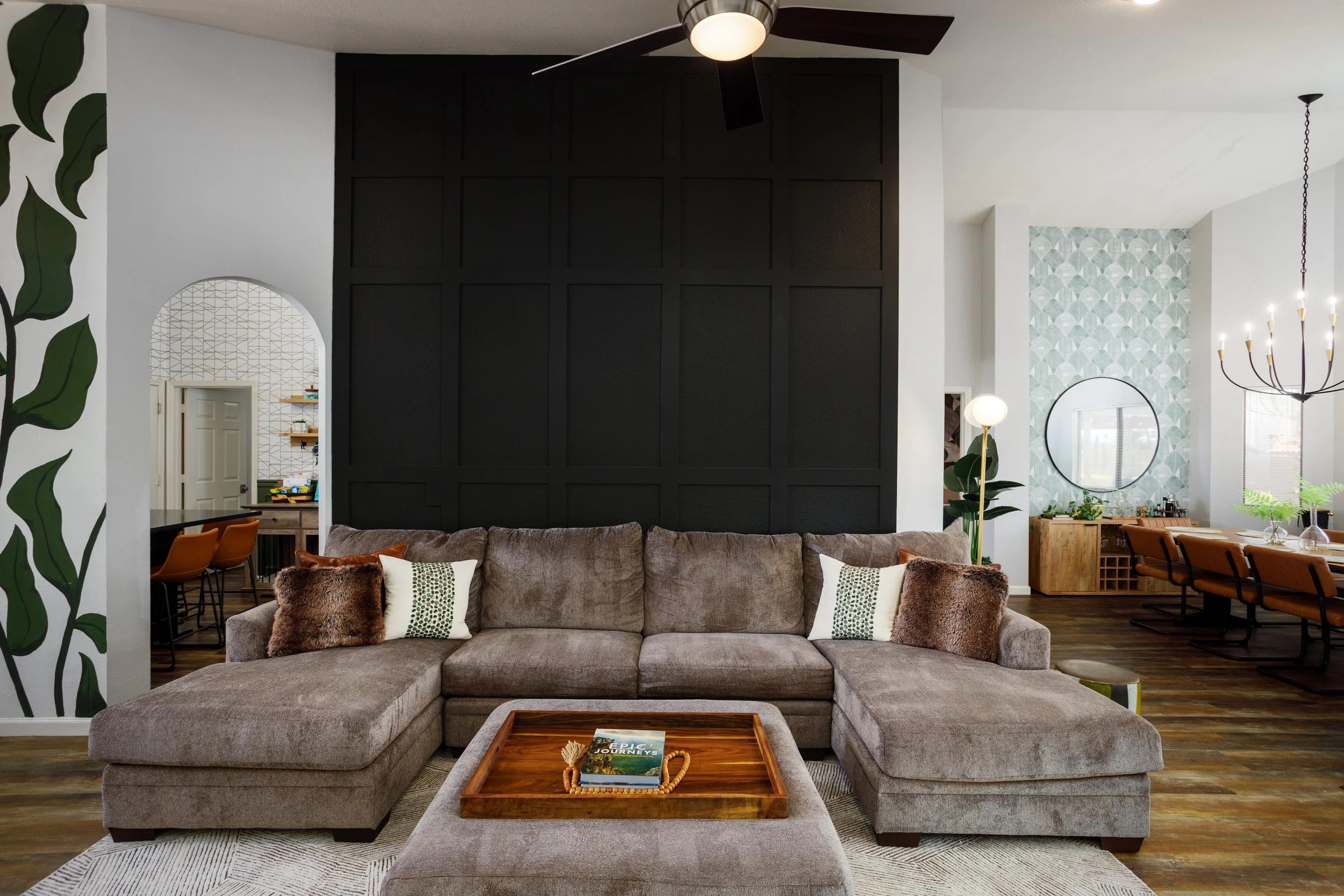 Living room with a large sectional gray sofa, decorative pillows, a wooden tray with a book on a coffee table, and a black accent wall with textured panels. In the background, there is a kitchen and dining area with orange chairs, a round mirror, a c