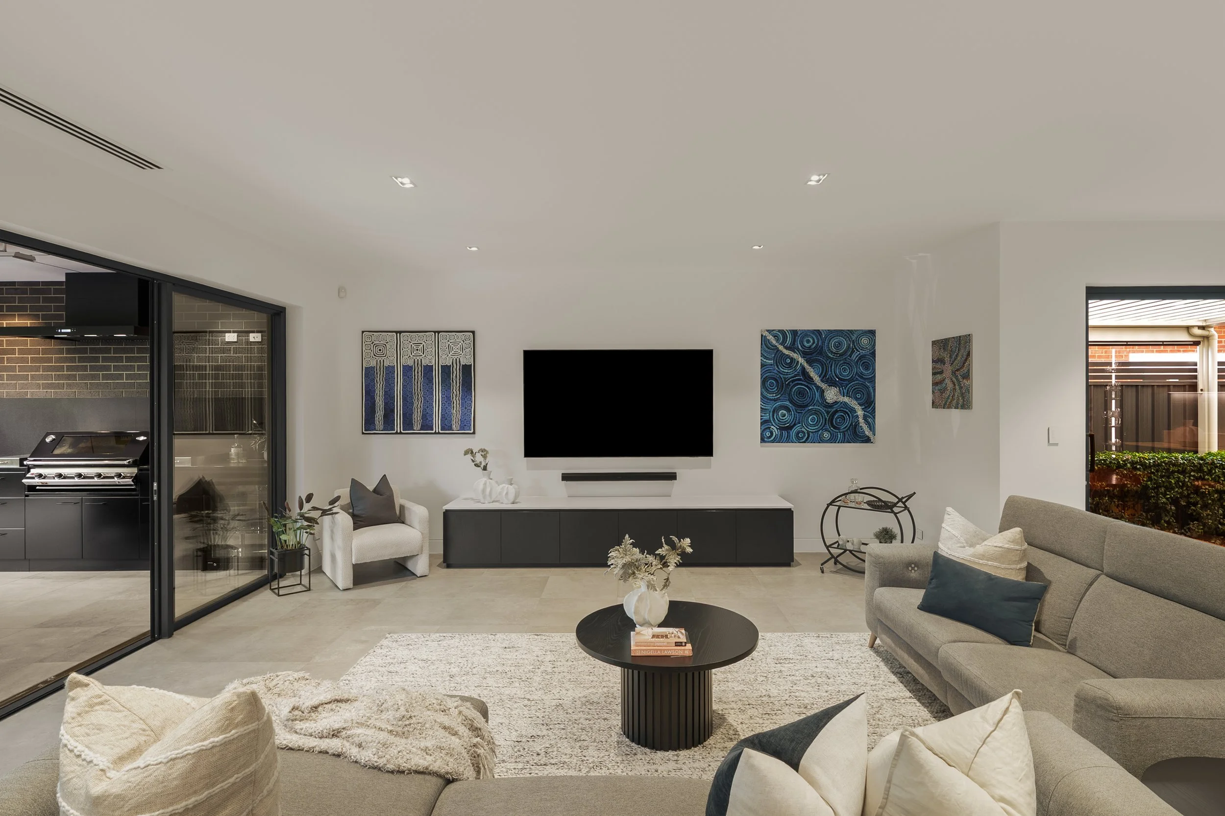 Modern living room with gray sofa, black round coffee table, abstract artwork on white walls, and large windows showing outdoor brick wall and plants.