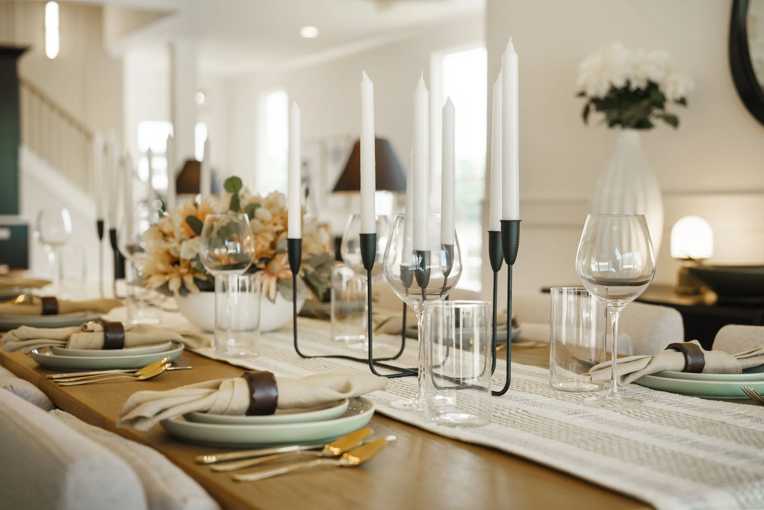 A dining table decorated with white candles in black candle holders, floral arrangements, wine glasses, and place settings with gold utensils and cream-colored napkins held by dark napkin rings in a bright, modern room.