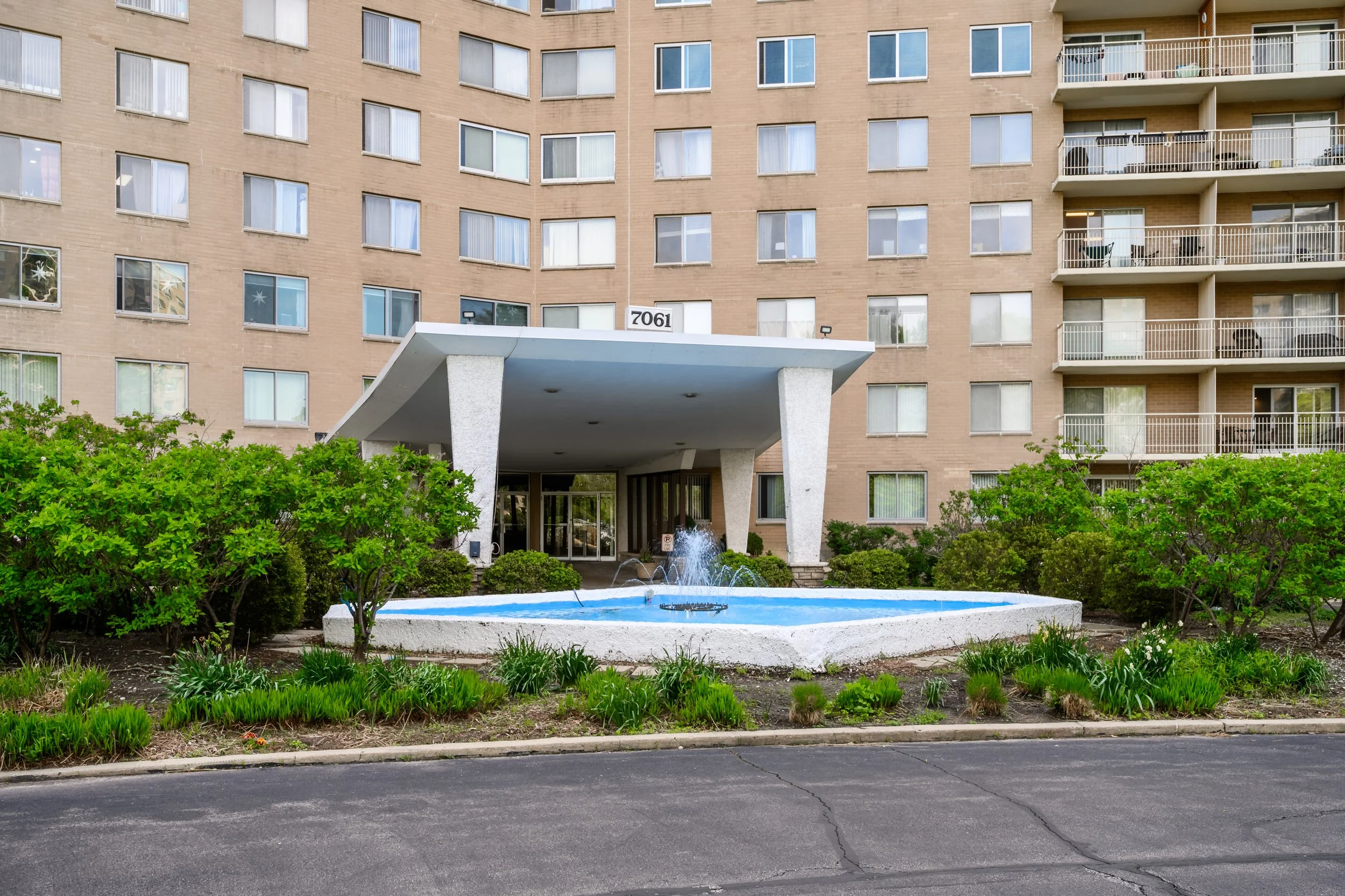 Front view of a residential apartment building with multiple floors and balconies. A covered entrance with a white canopy and the number 7061 above it. In front, there are green bushes, a small fountain, and a street with a curb.