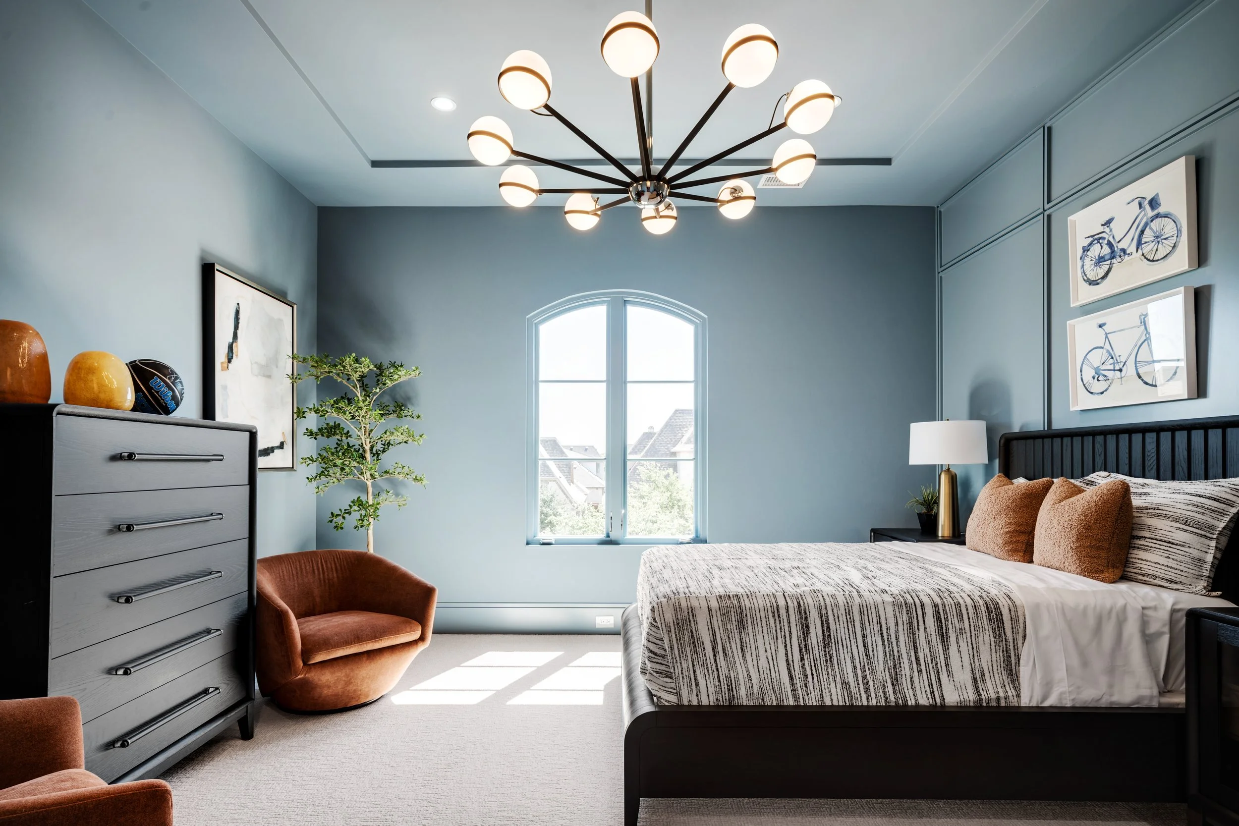 A modern bedroom with a large window, blue walls, and a stylish chandelier. The bed has a striped duvet and two orange pillows, with a side table and lamp. There's a dresser with decorative vases, a framed artwork, and a potted plant in the room.